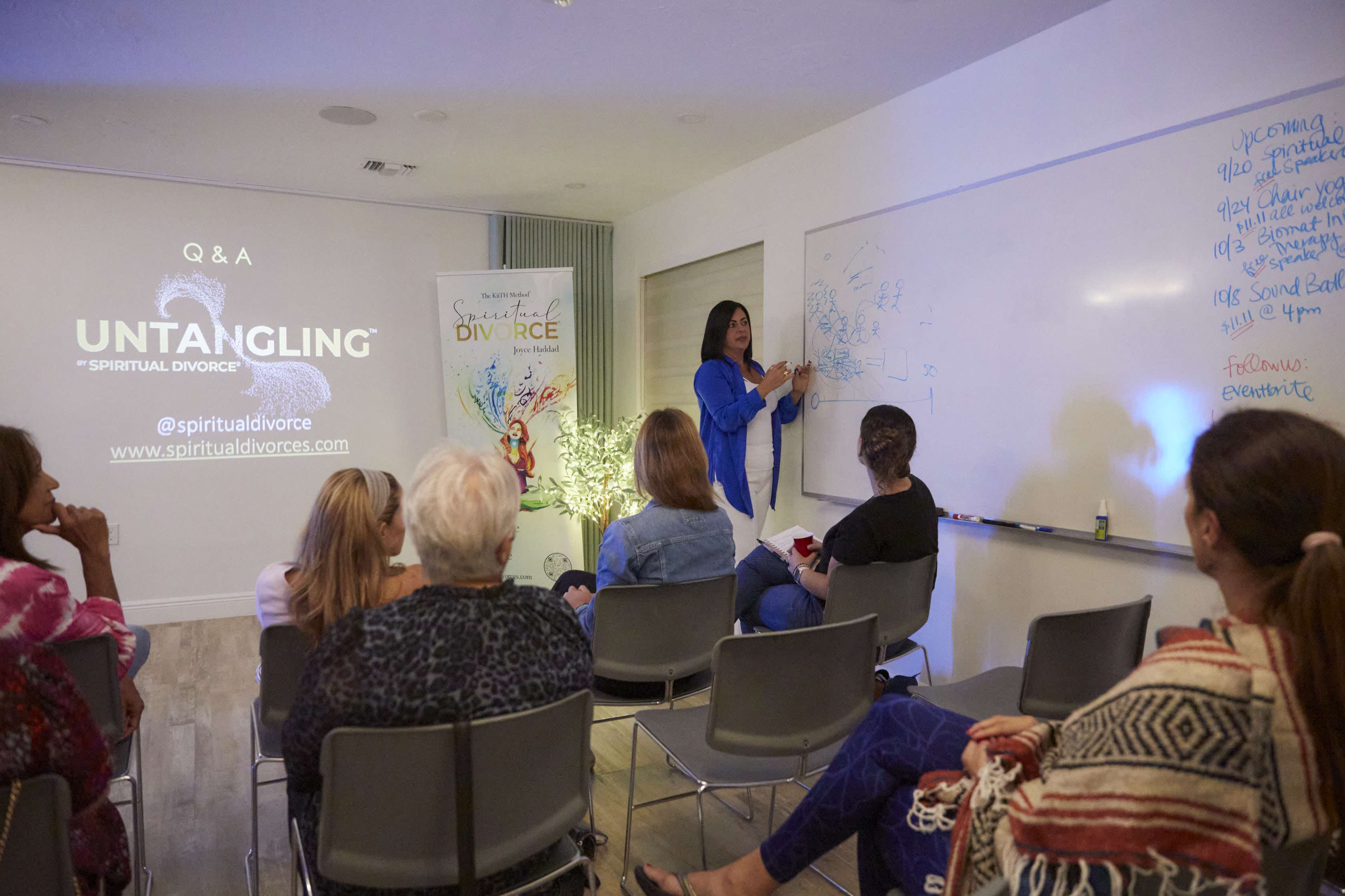 A woman is presenting a diagram on a whiteboard during a Q&A session in a brightly lit room with an audience seated in chairs.