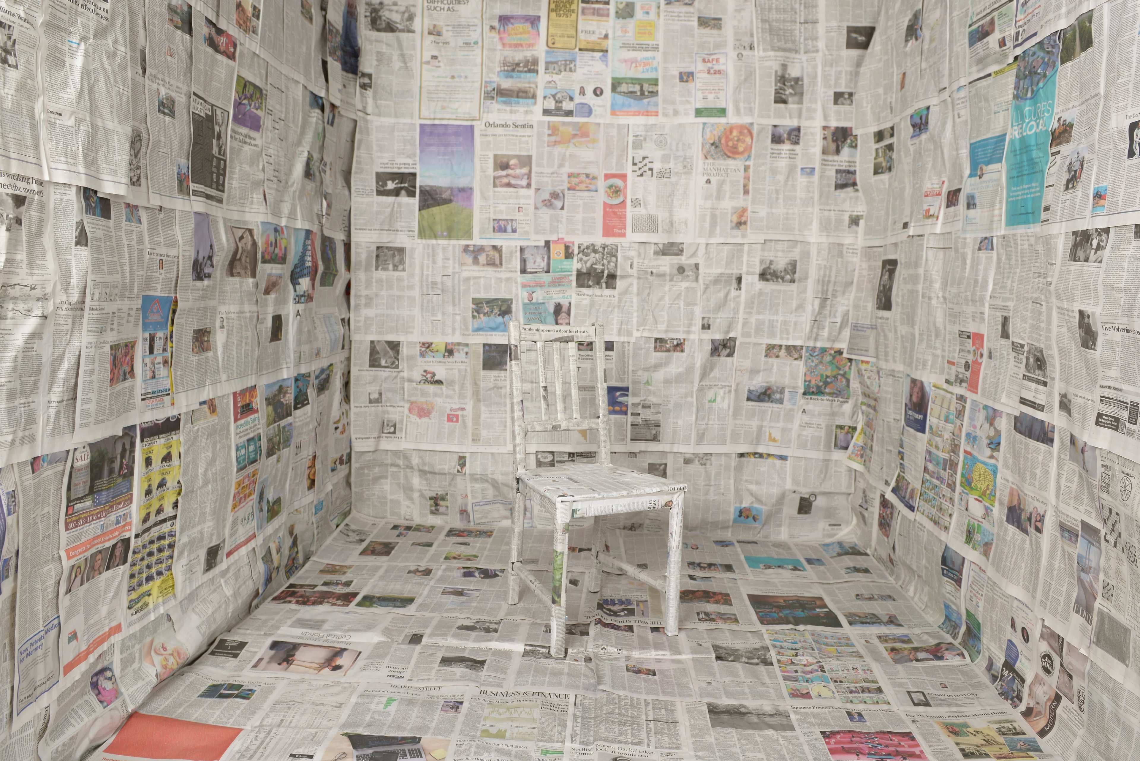 A single white chair is placed in the center of a room where the walls and floor are covered entirely in newspaper.
