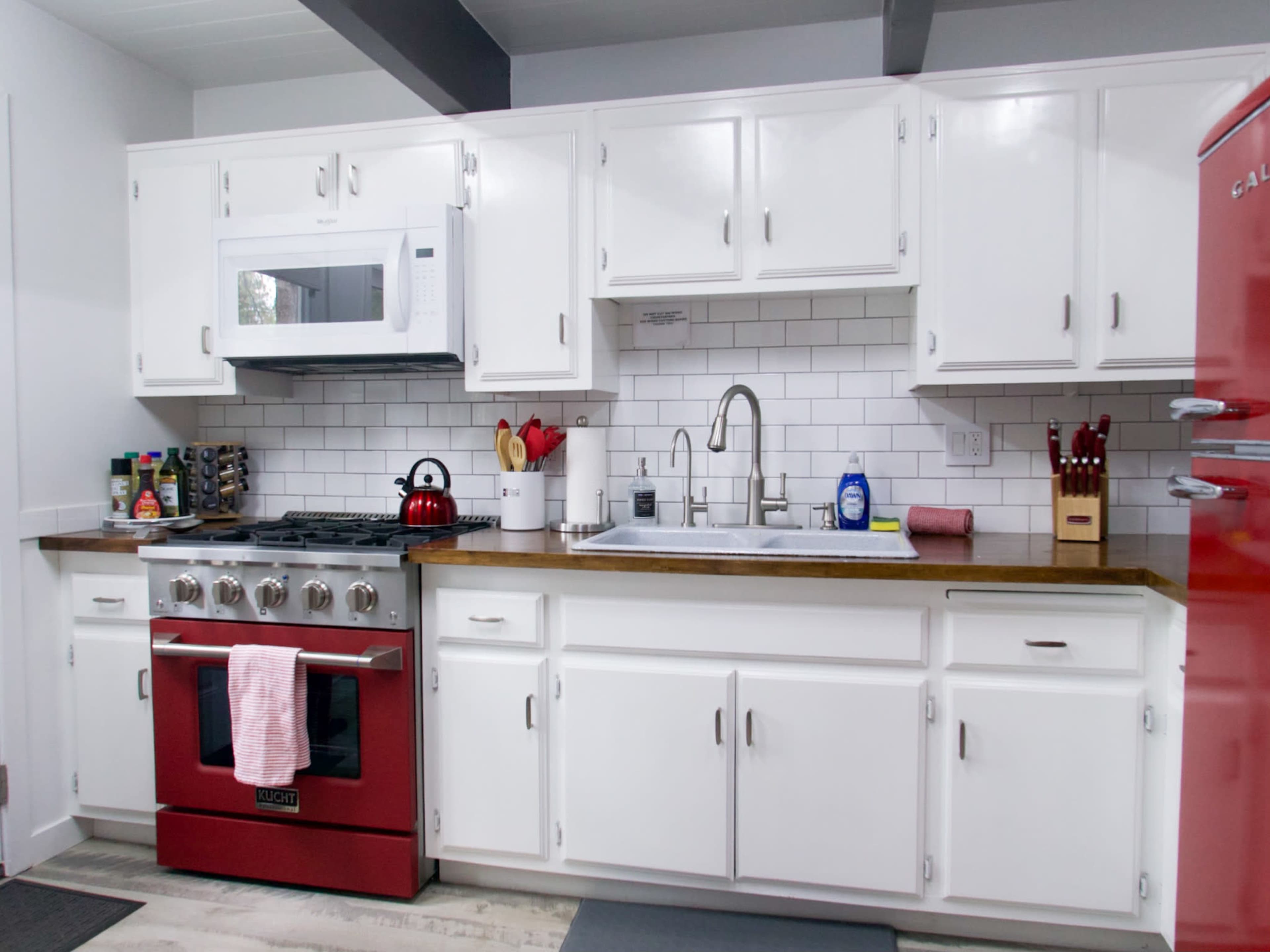 The image shows a modern kitchen featuring white cabinets, a stainless steel sink, a red stove, and a microwave above the counter, with a backsplash of white tiles.