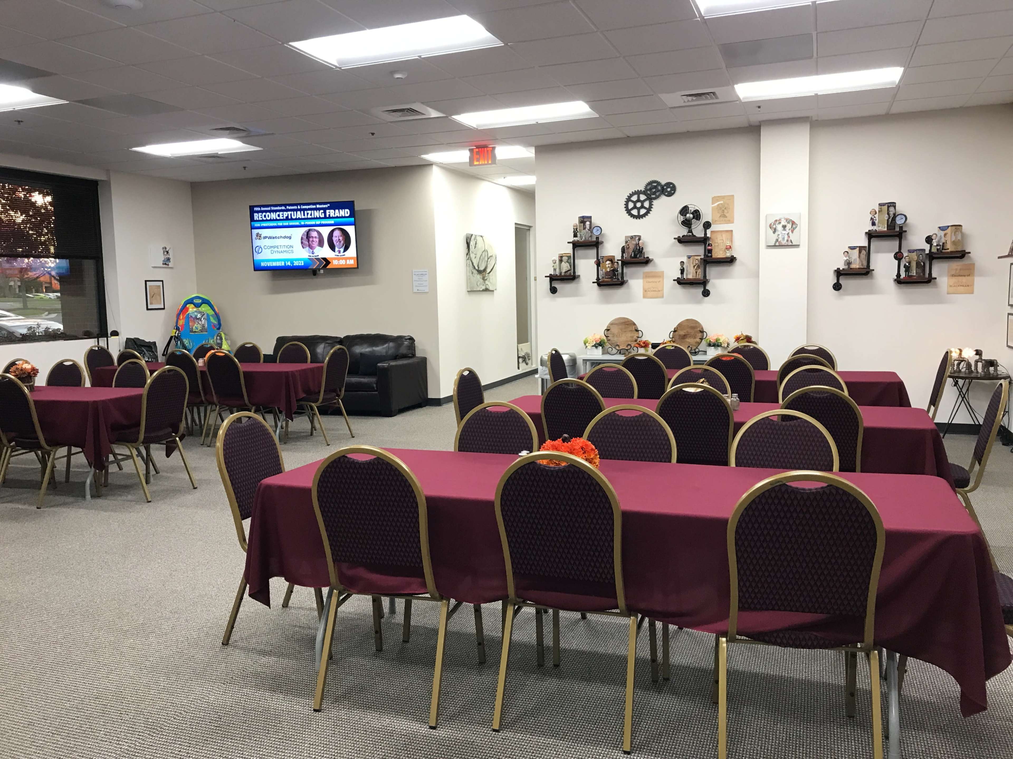 A spacious room features several round tables with maroon tablecloths and chairs, alongside a wall displaying decorative shelves and a screen showing information.