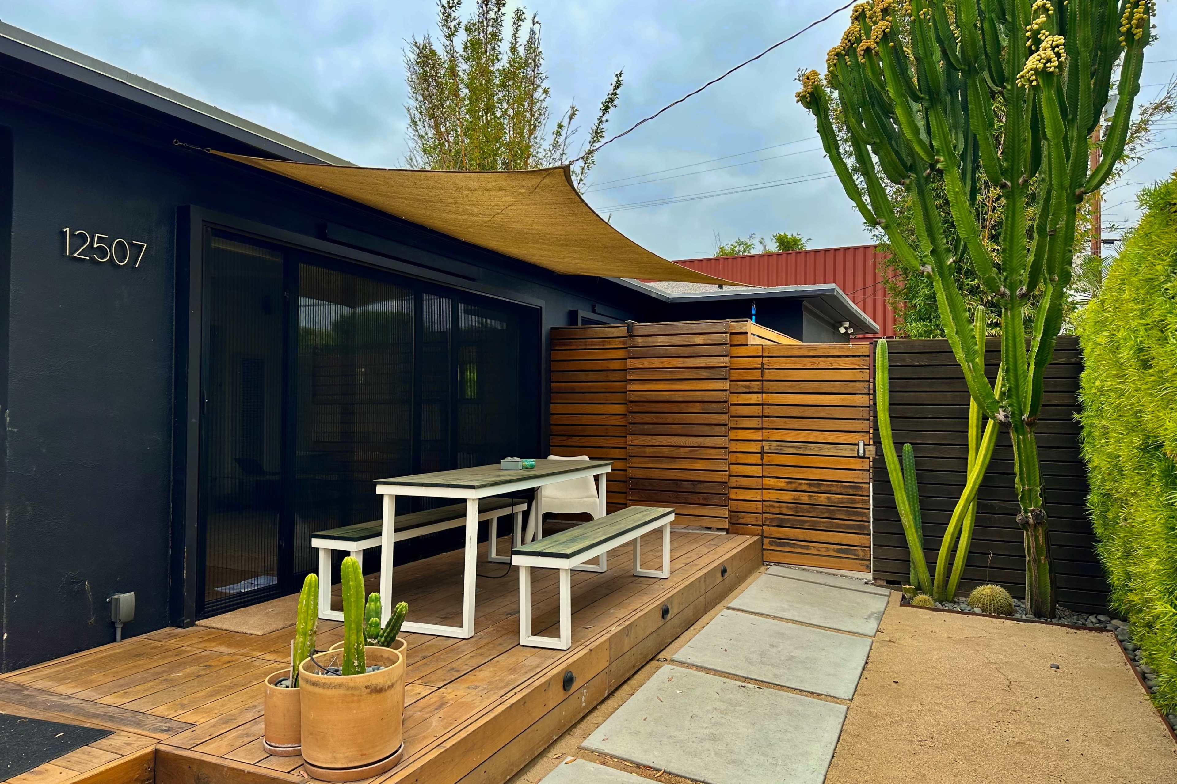 The image shows a modern outdoor space with a wooden deck, a dining table and benches, a shade sail overhead, and tall cacti along the perimeter.