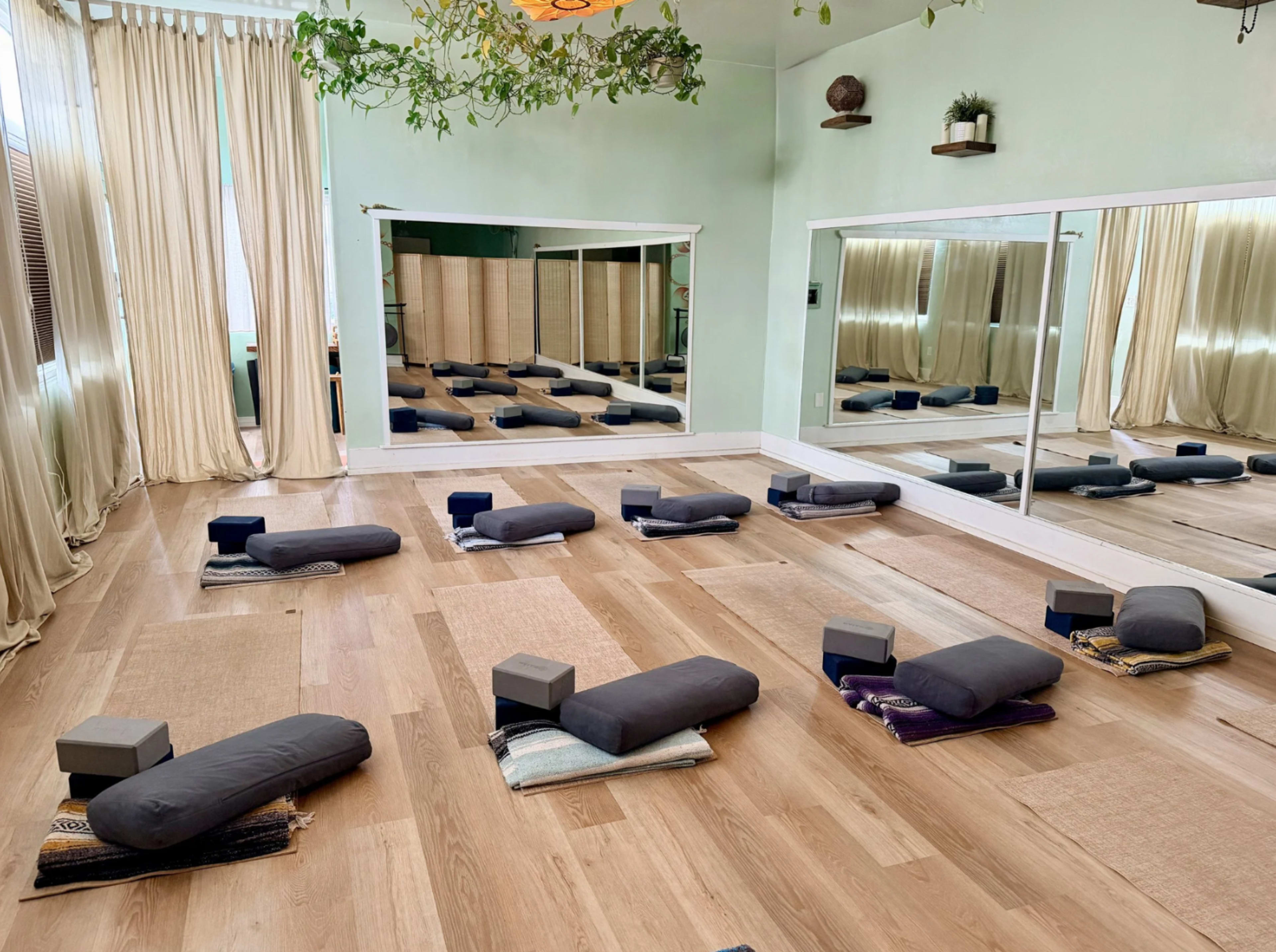 A yoga studio features several mats and bolsters arranged neatly on a wooden floor, with mirrors and natural light enhancing the serene atmosphere.