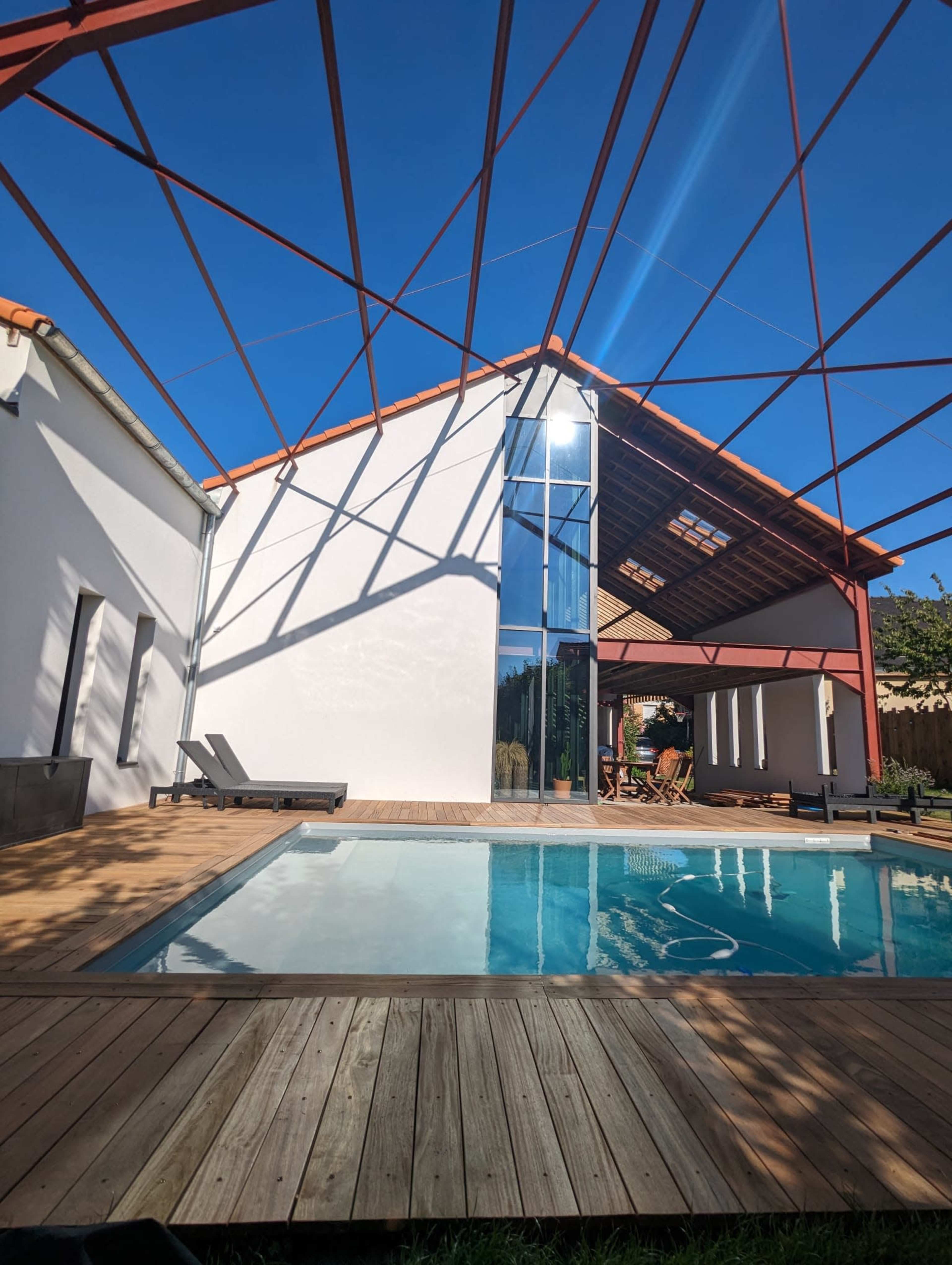 A modern building with a large glass facade, a swimming pool in the foreground, and a wooden deck under a clear blue sky with a steel frame structure overhead.