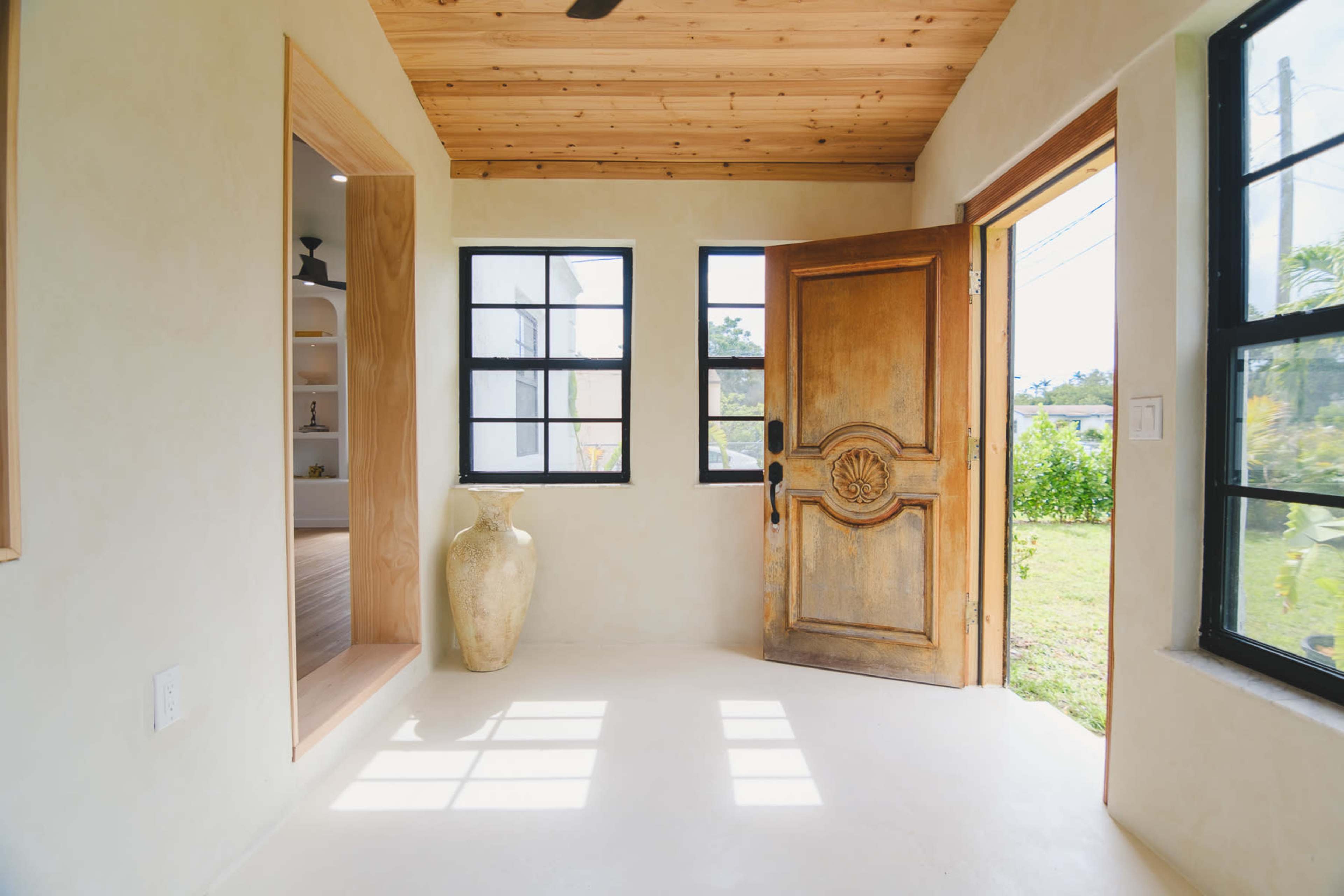 The image shows a bright entryway with a wooden door partially open, revealing a view of greenery outside.