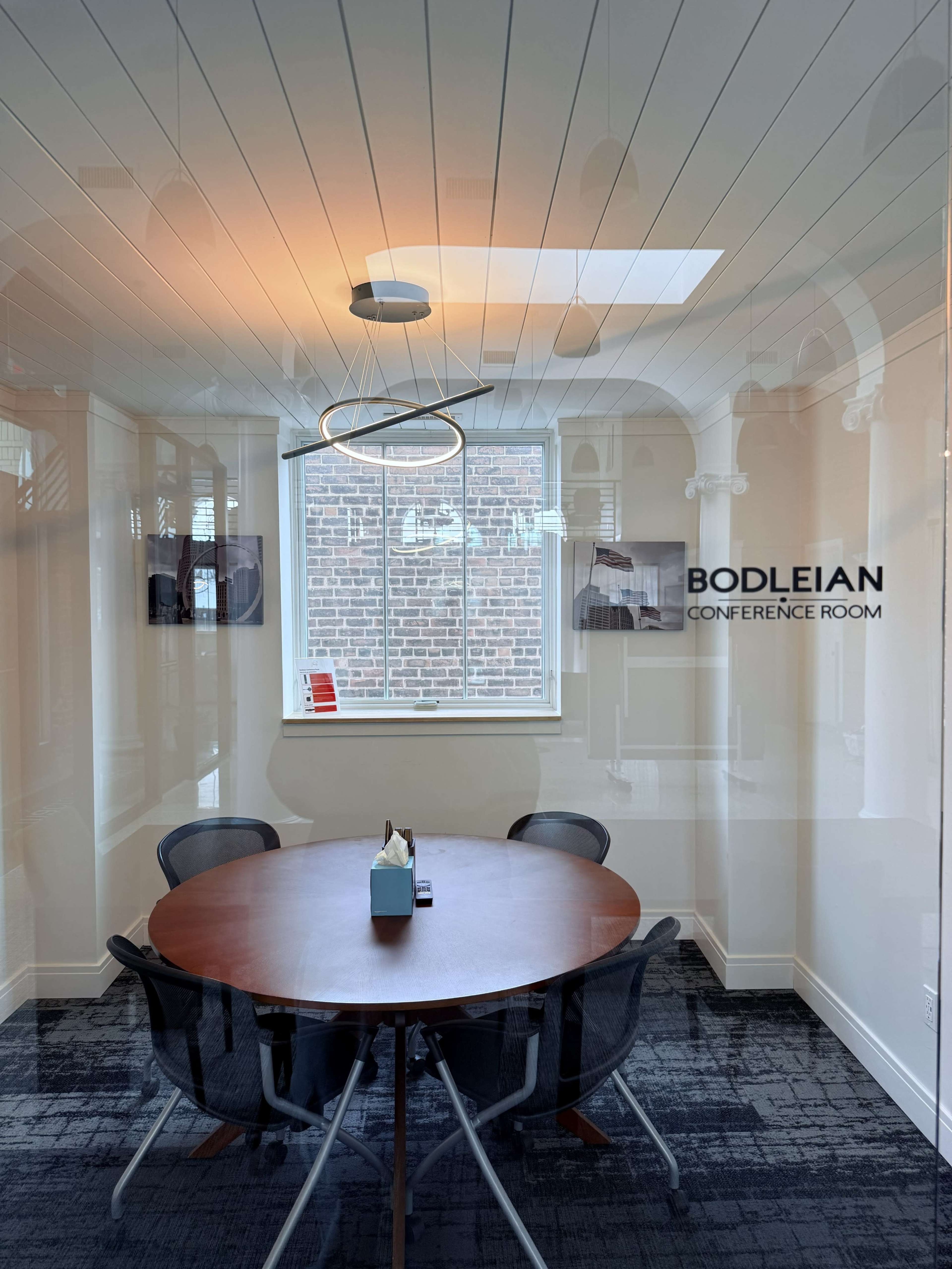 A circular wooden conference table surrounded by four chairs is set in a conference room with large glass windows.