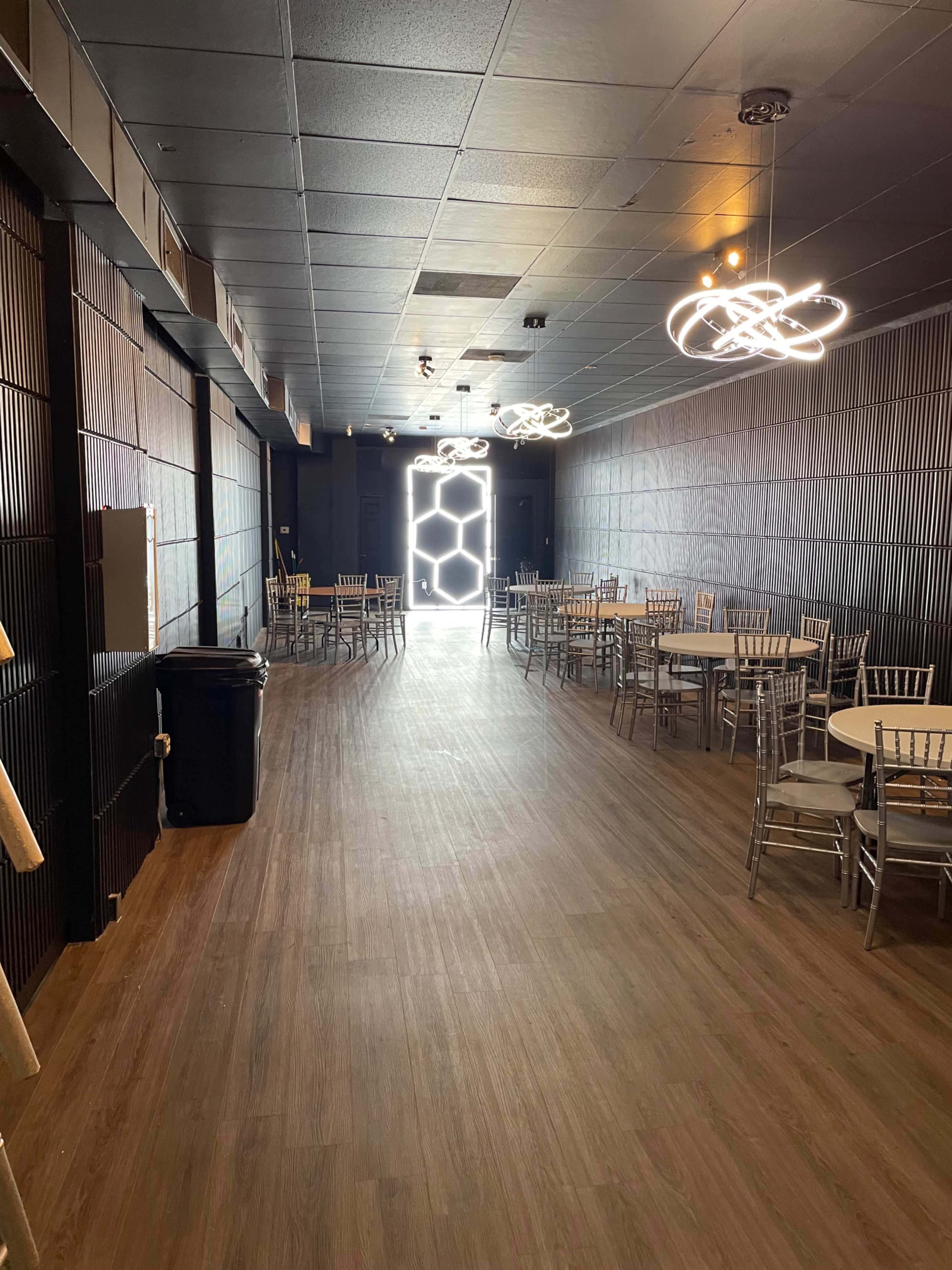 A spacious, dimly lit room with wooden flooring, round tables surrounded by metal chairs, and modern light fixtures hanging from the ceiling.