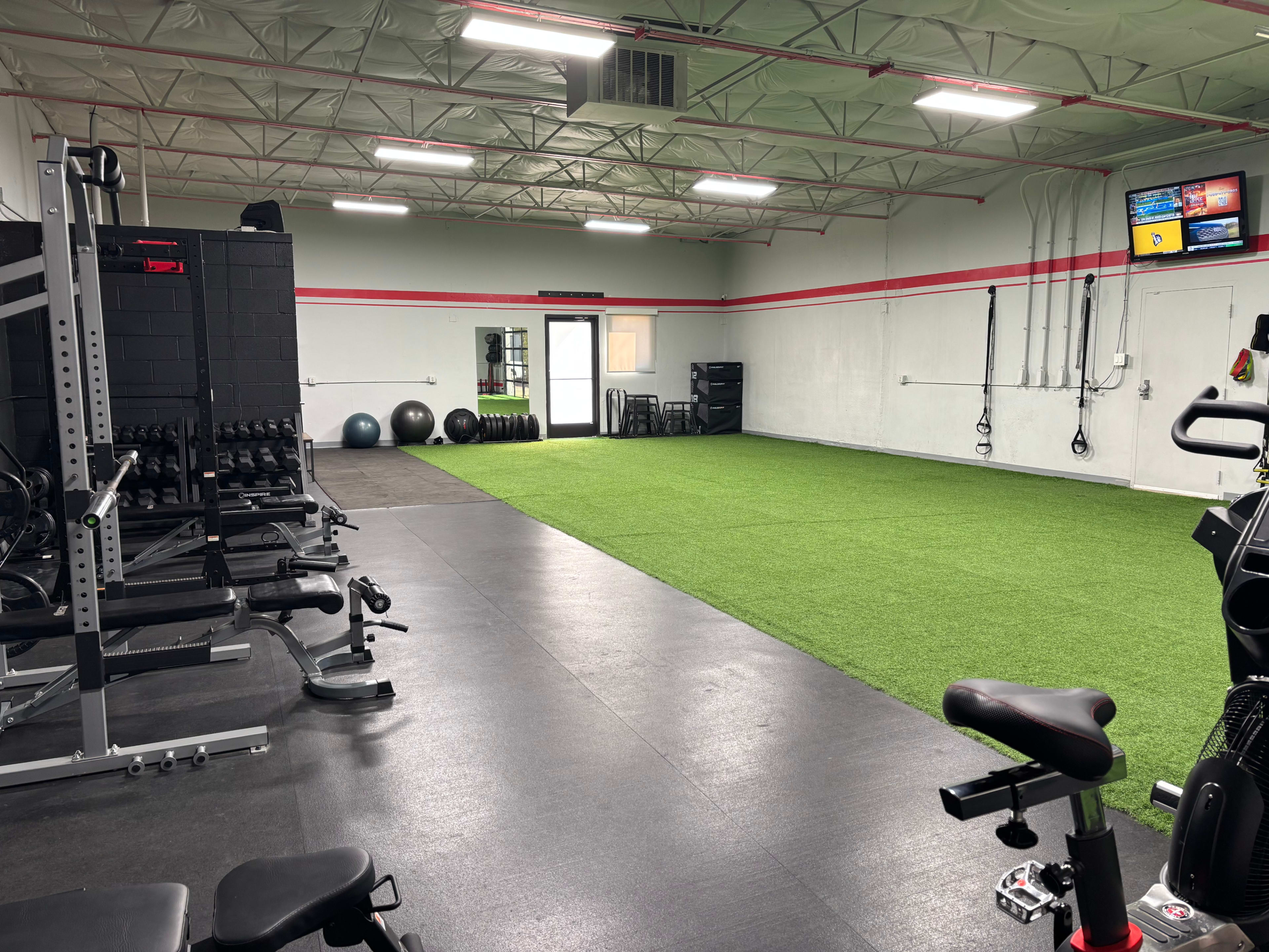The image shows an indoor gym with exercise equipment on one side and a turf area on the other, featuring weights, stability balls, and various fitness machines.