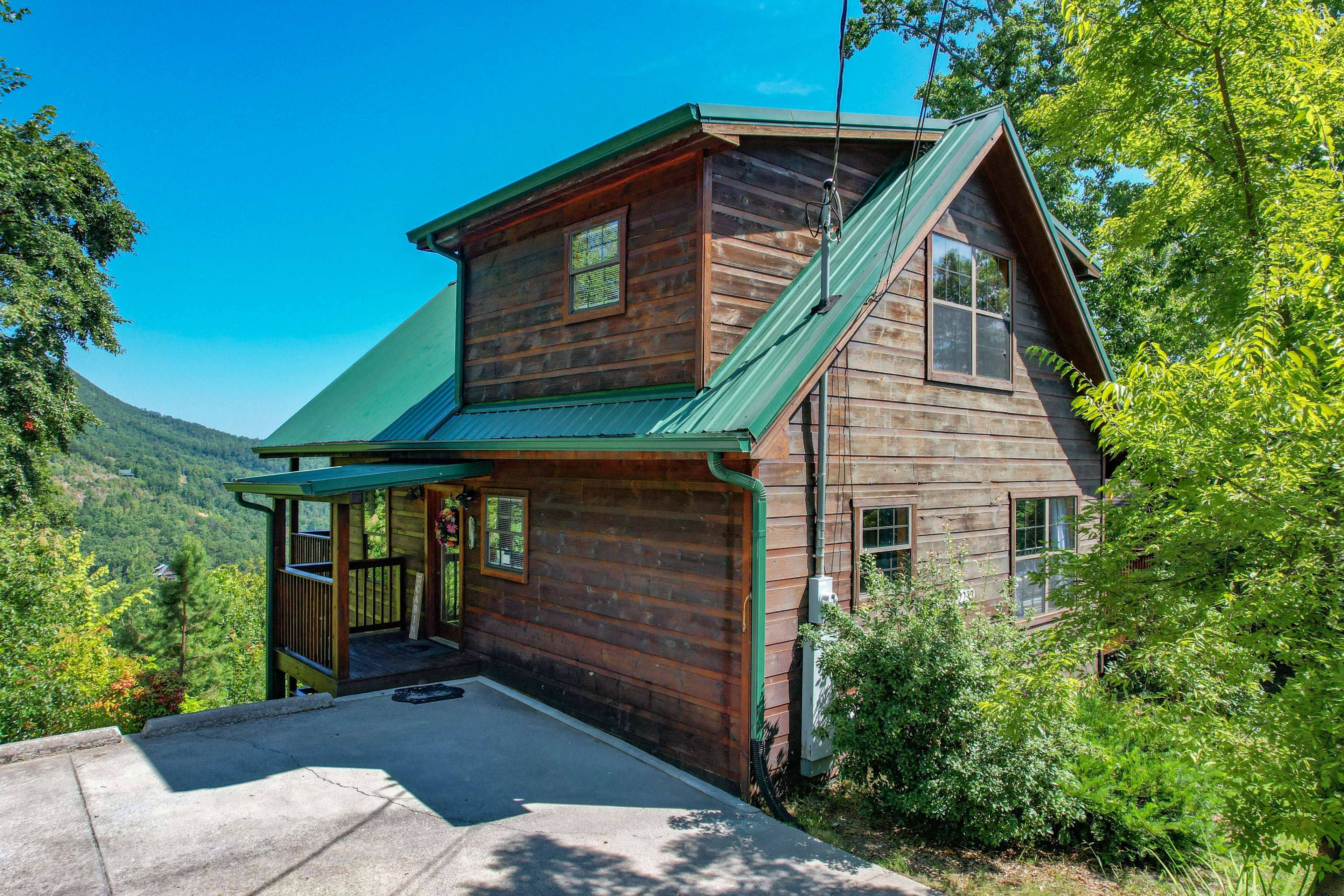 A wooden house with a green metal roof is situated on a slope surrounded by trees and mountains.