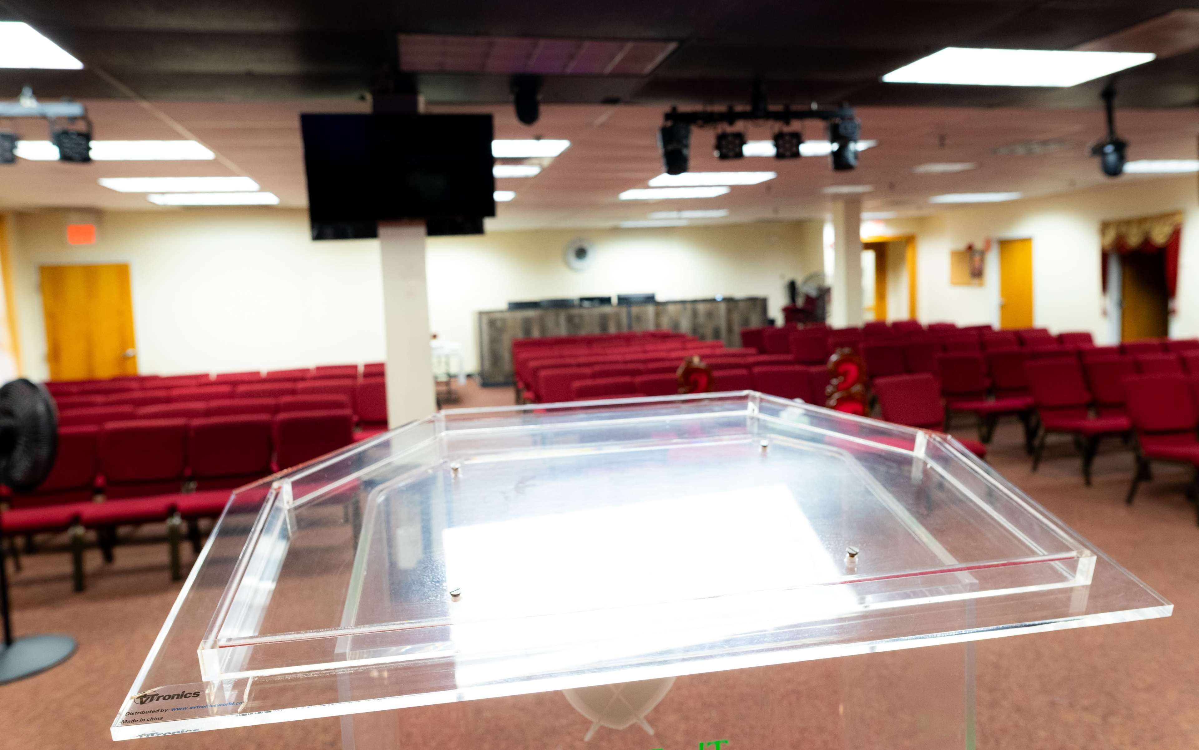 A clear lectern in the foreground, with rows of red chairs arranged in a semi-circle in a well-lit auditorium space.