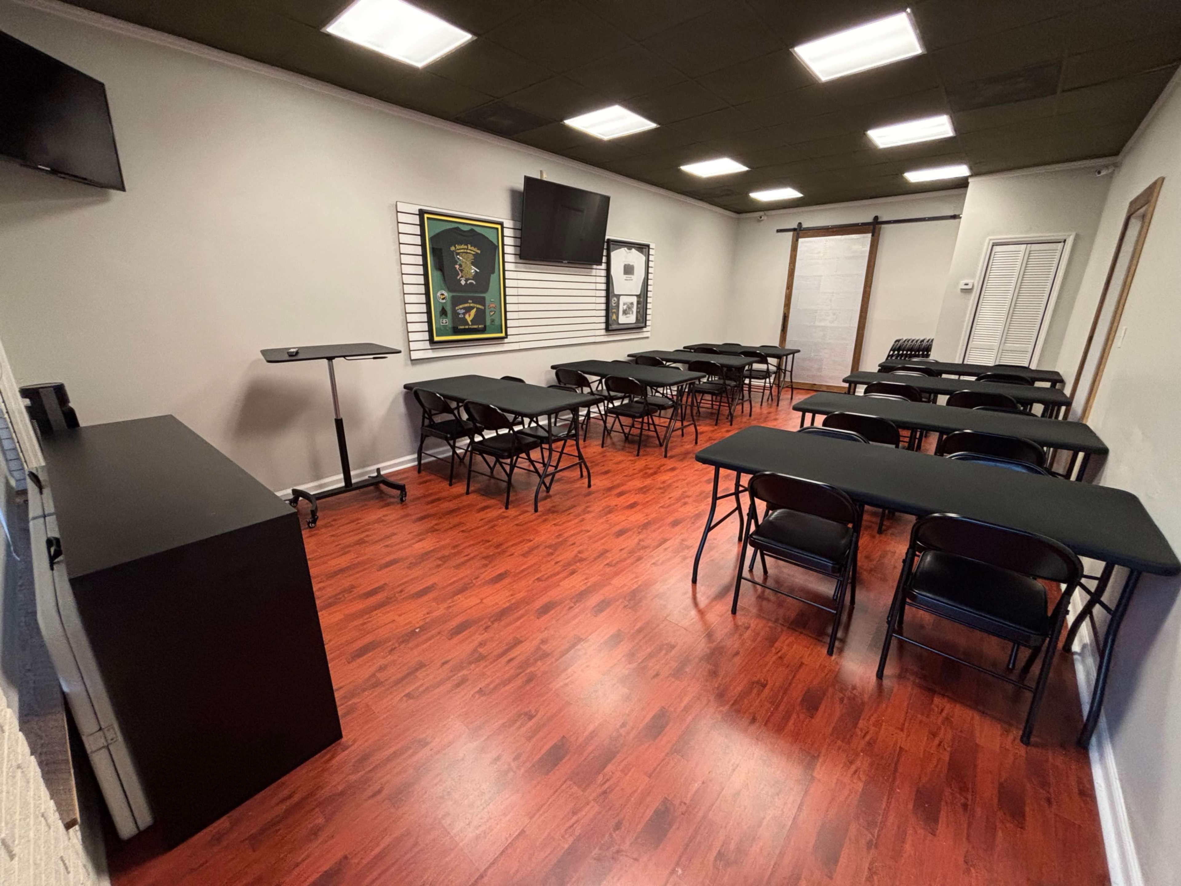 The image shows a room with several black tables and chairs arranged for a meeting or event, along with a television and framed artwork on the walls.