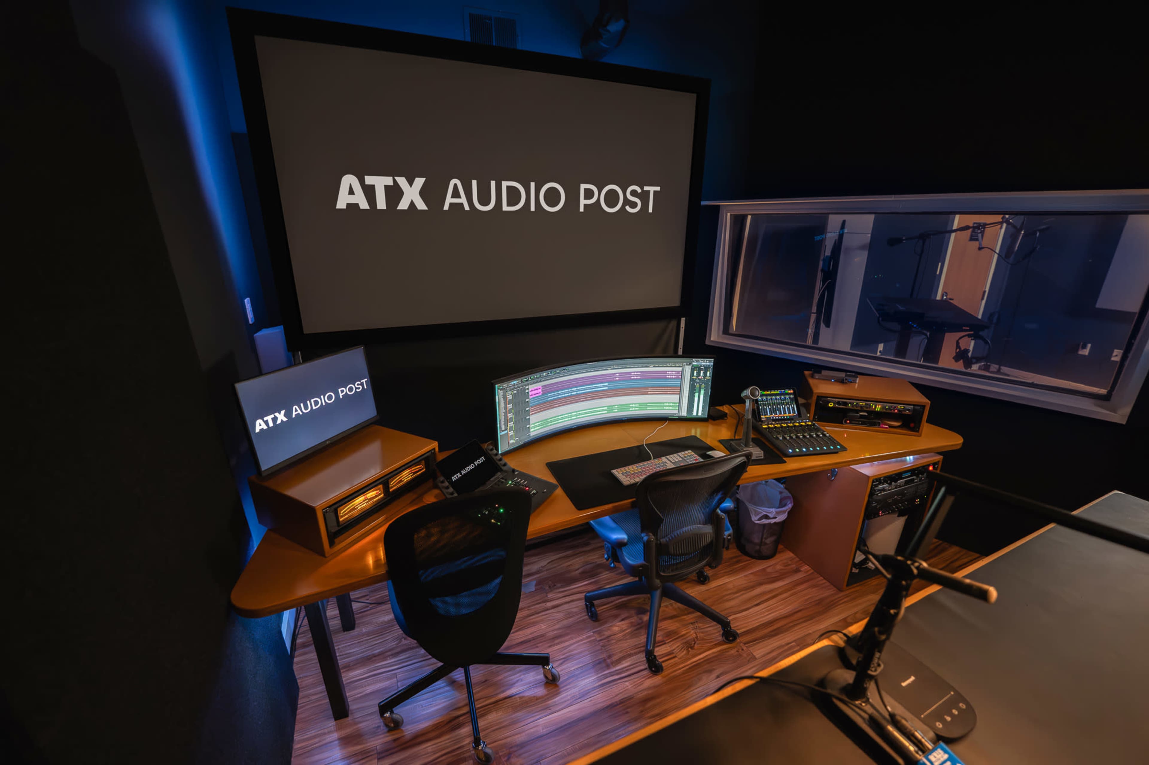 The image shows a modern audio post-production studio with a large screen displaying "ATX AUDIO POST," a control desk, and various audio equipment set up around a chair.