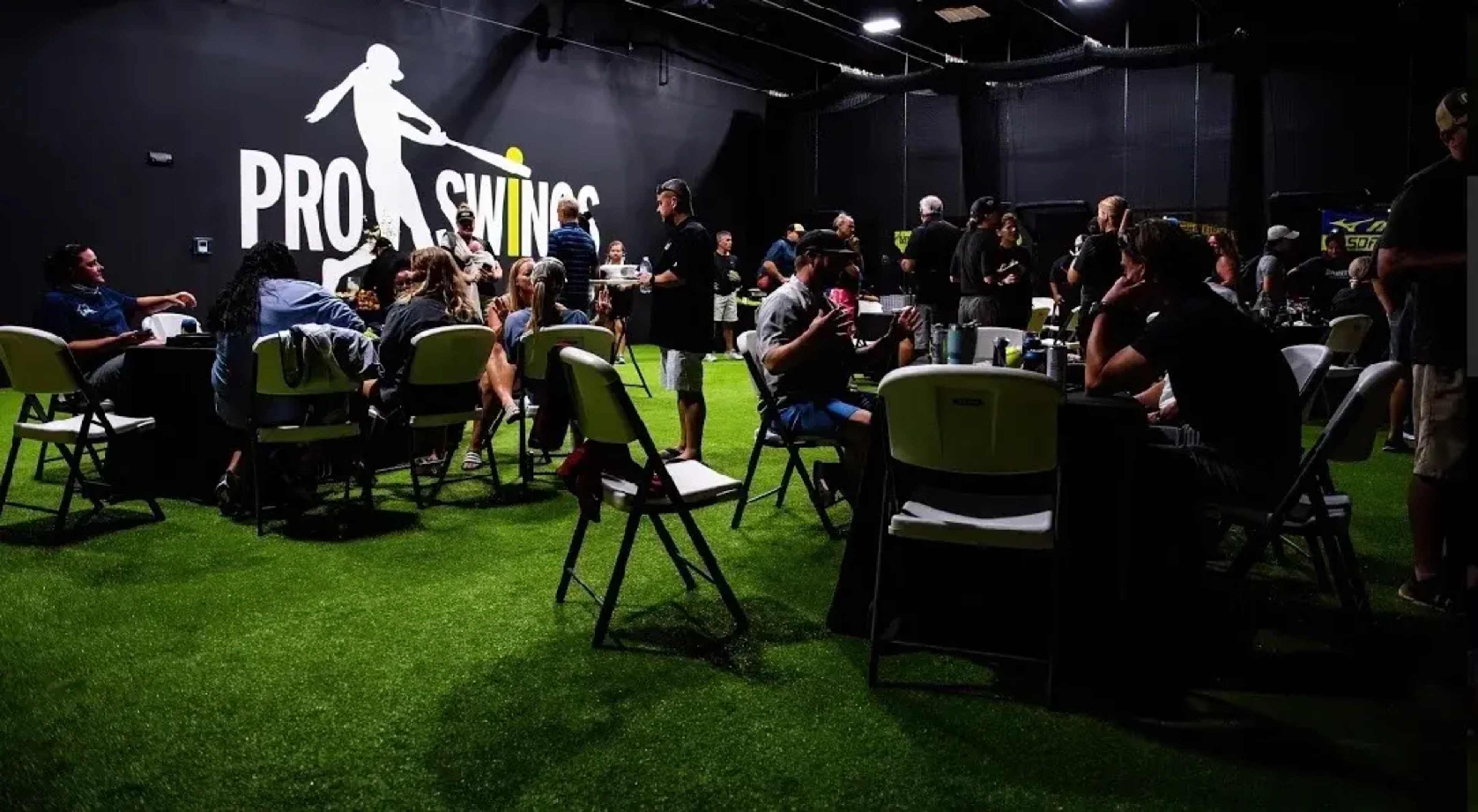 A group of people is gathered indoors at a facility called Pro Swings, engaging in various activities around tables on artificial grass.