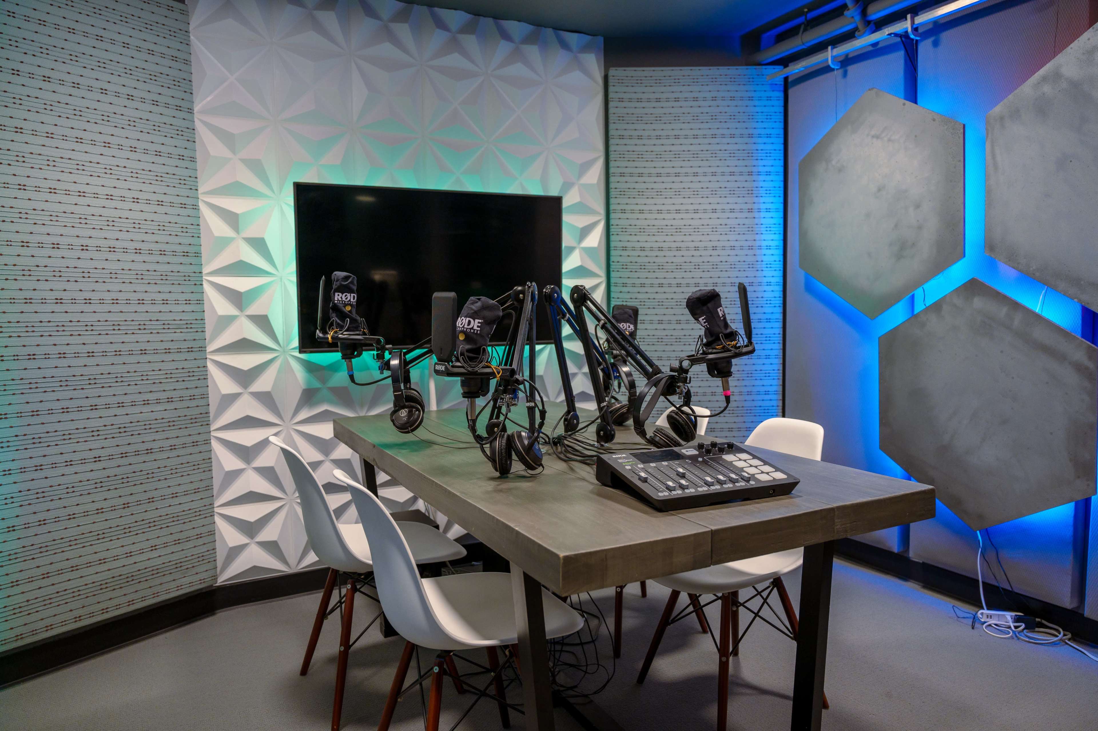 A modern podcast recording studio with a table, several microphones on adjustable arms, a control panel, and a television screen mounted on the wall.