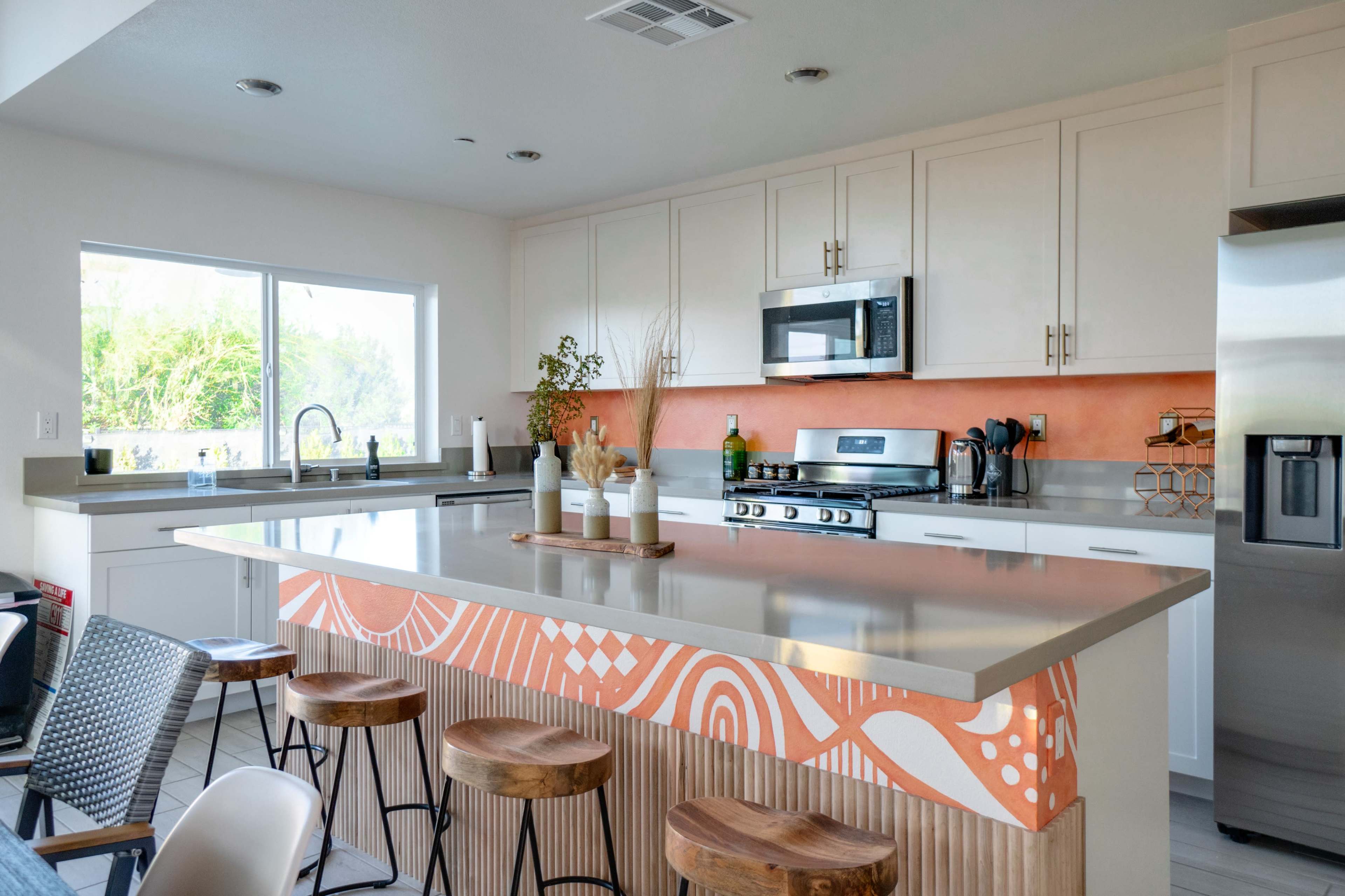 A modern kitchen features a large island with wooden stools, stainless steel appliances, and a bright orange accent wall.