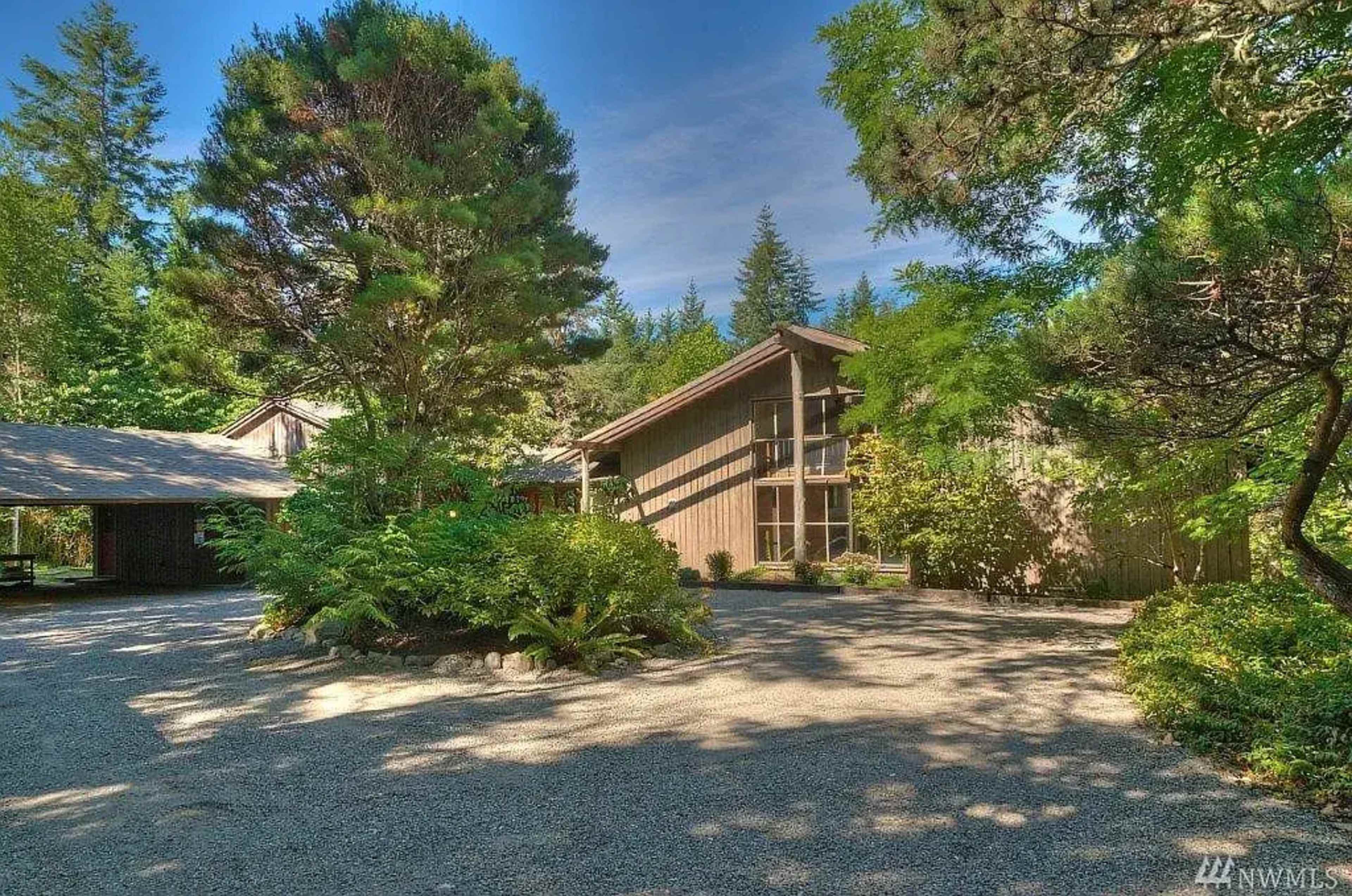 A modern, two-story wooden house is surrounded by lush greenery and gravel pathways under a clear blue sky.