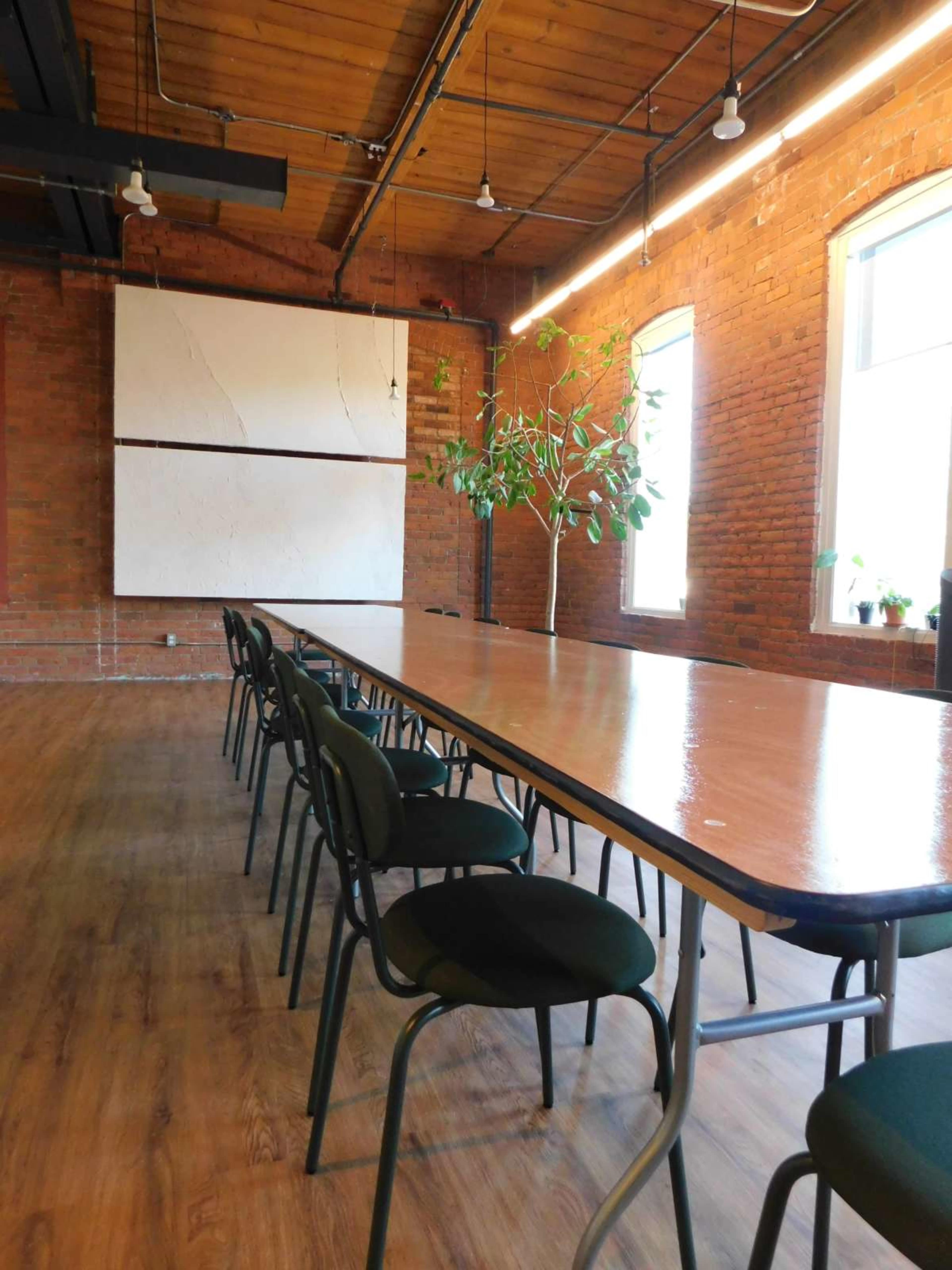 A long wooden table with chairs is set up in a room featuring brick walls and large windows, with a plant beside the table and a blank wall surface at the end.