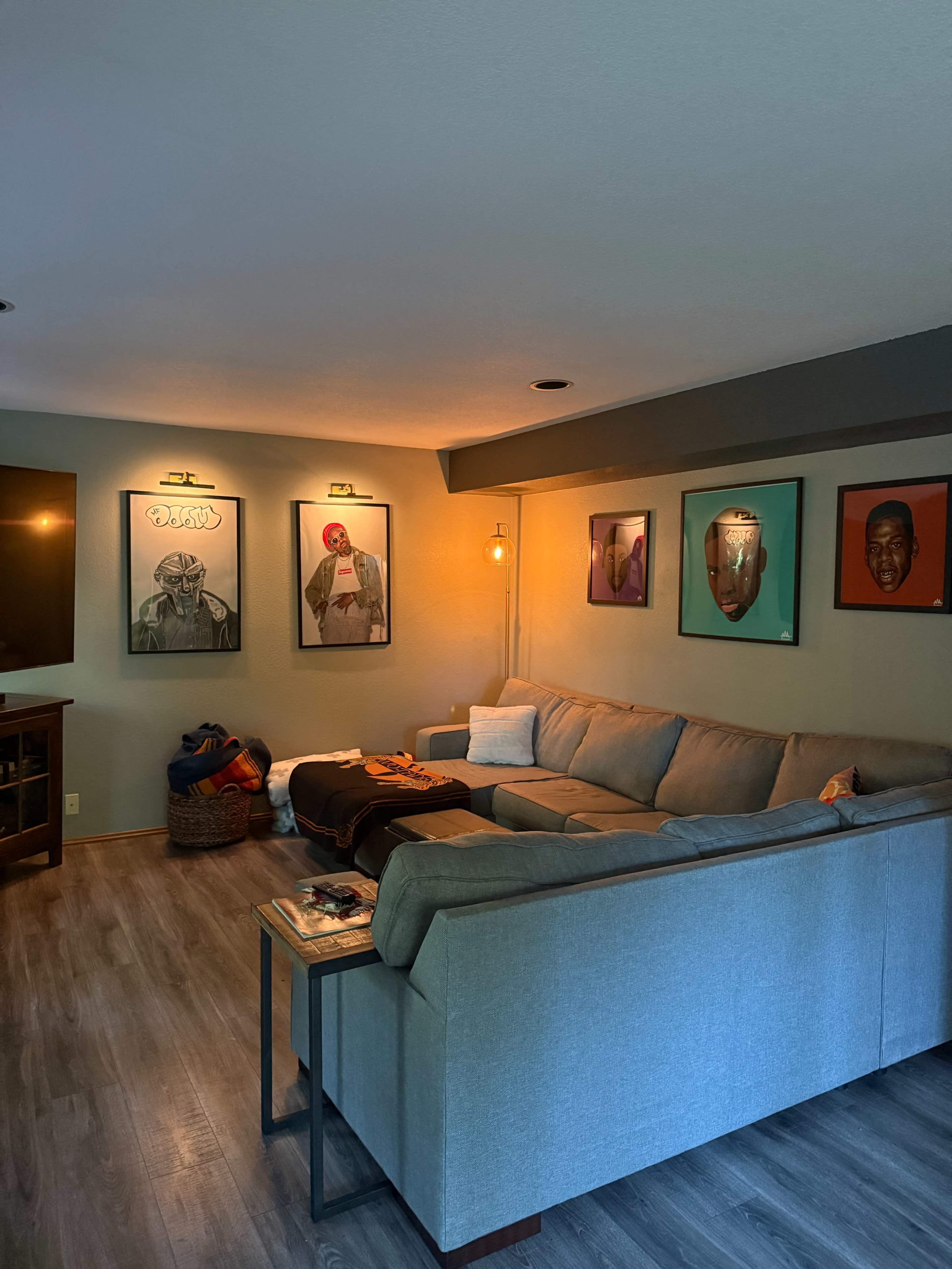 The image shows a cozy living room featuring a light gray sectional sofa, a wooden side table, and several framed portraits on the wall, illuminated by warm overhead lighting.