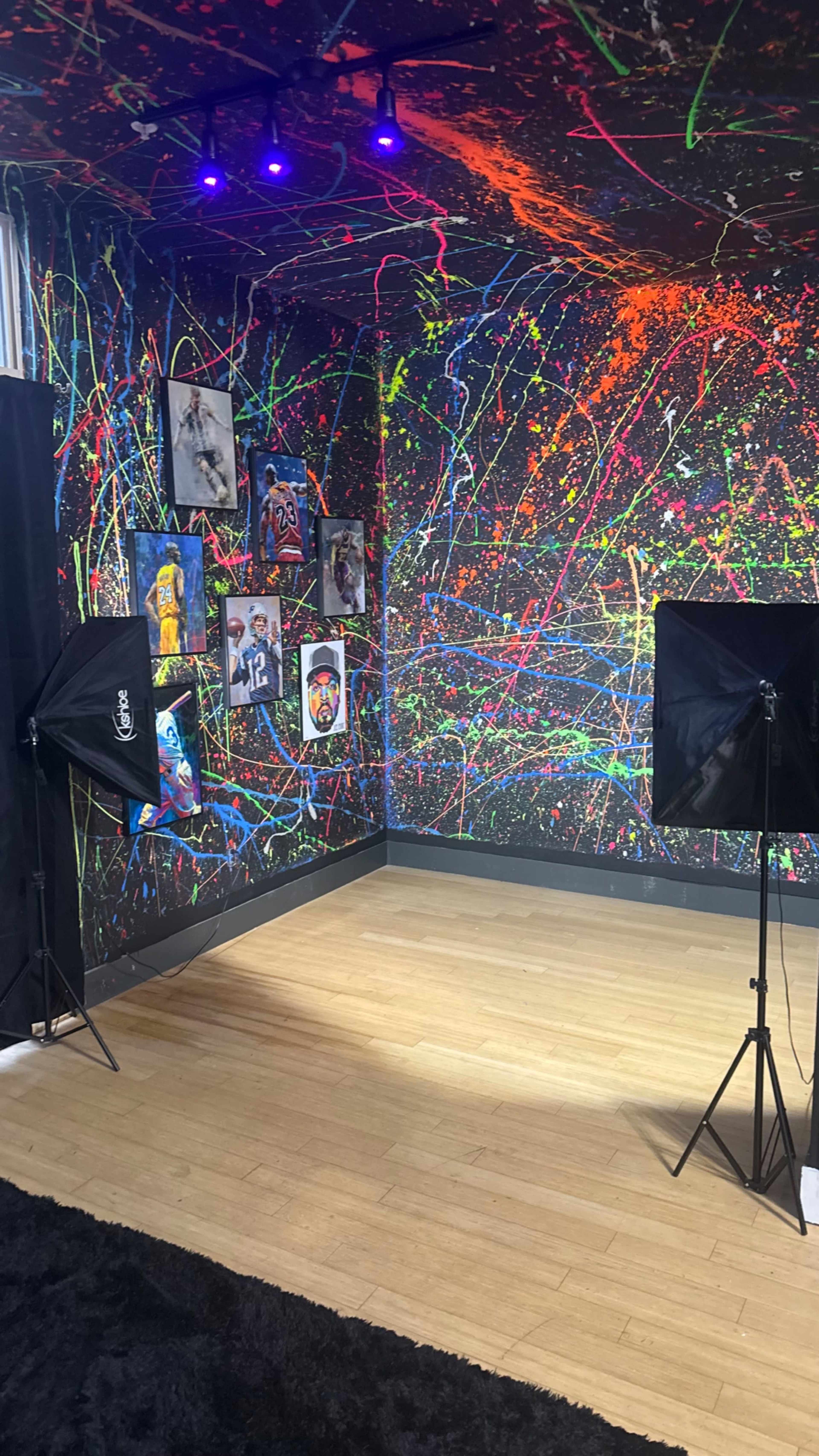 The image shows a brightly colored room with a splatter-painted wall, showcasing framed artwork, and equipped with two photography lights on tripods.