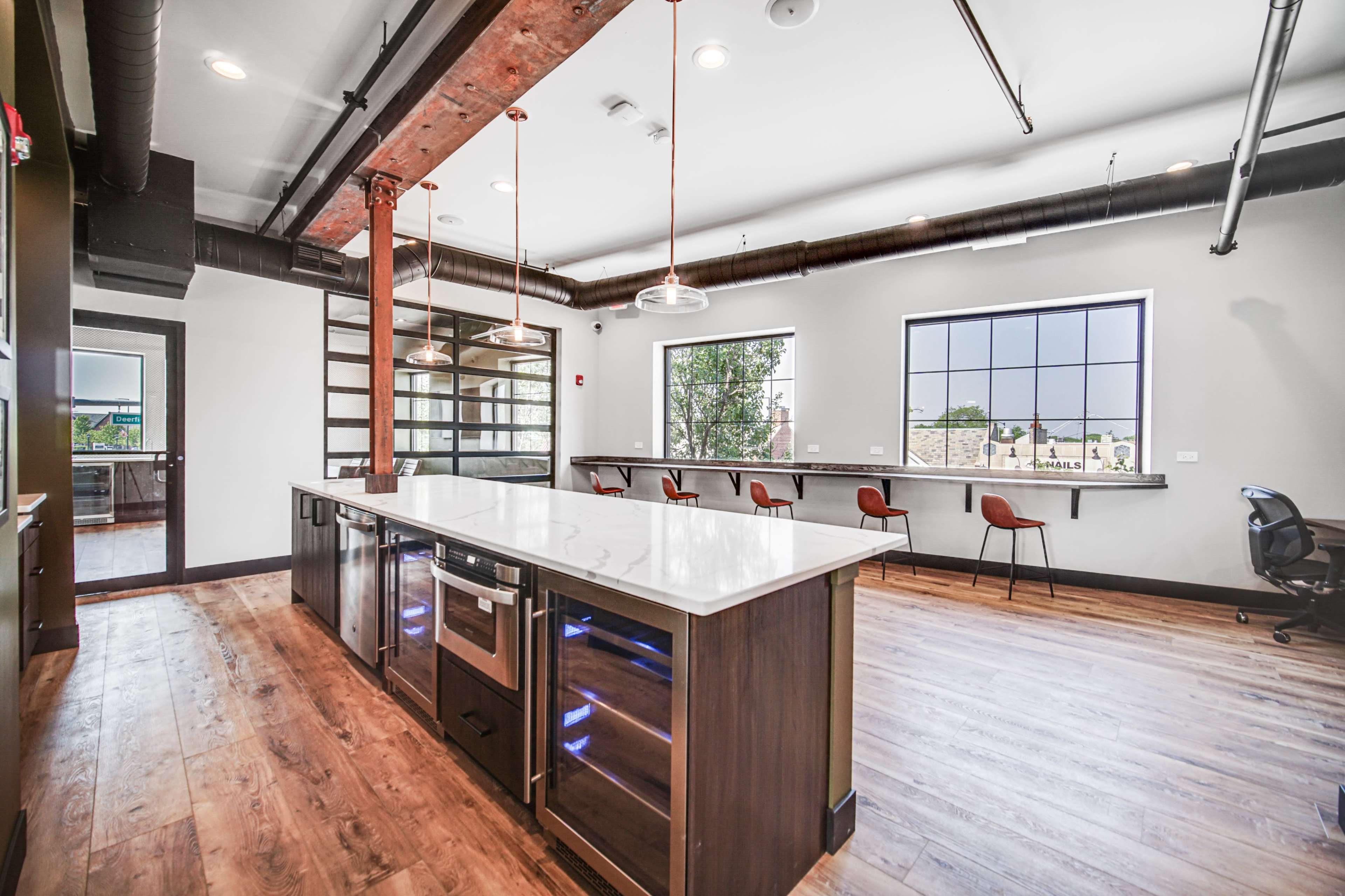 A modern kitchen features a large island with appliances and a row of bar stools along one wall, illuminated by pendant lights and complemented by large windows.