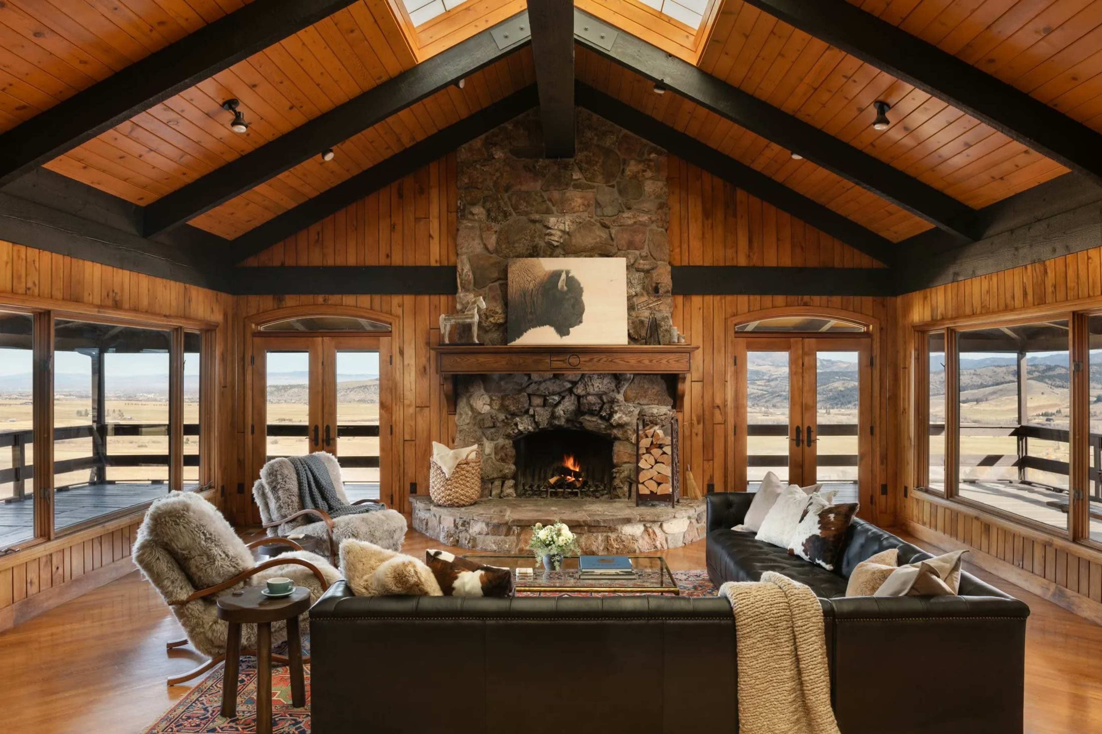 The image shows a cozy living room with a stone fireplace, wooden beams, and large windows overlooking a scenic landscape.