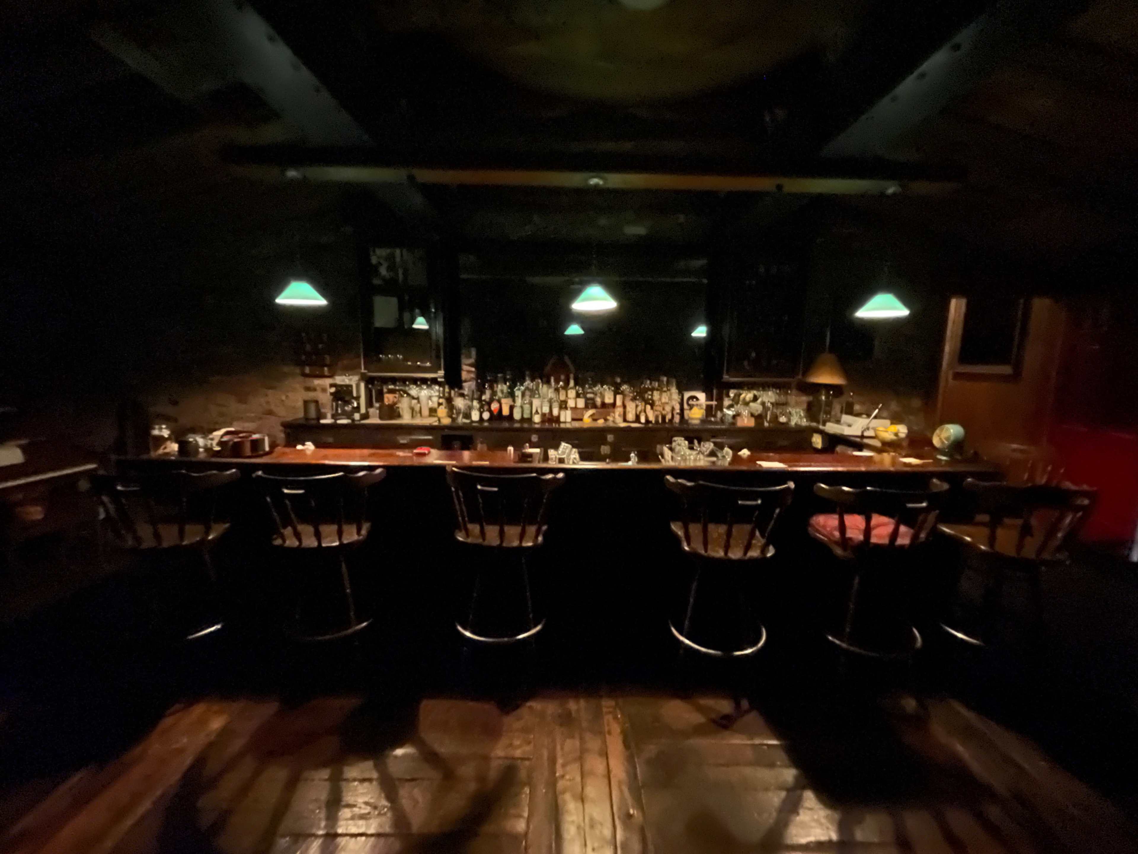 A dimly lit bar with a long wooden counter, several stools, and shelves lined with various bottles.