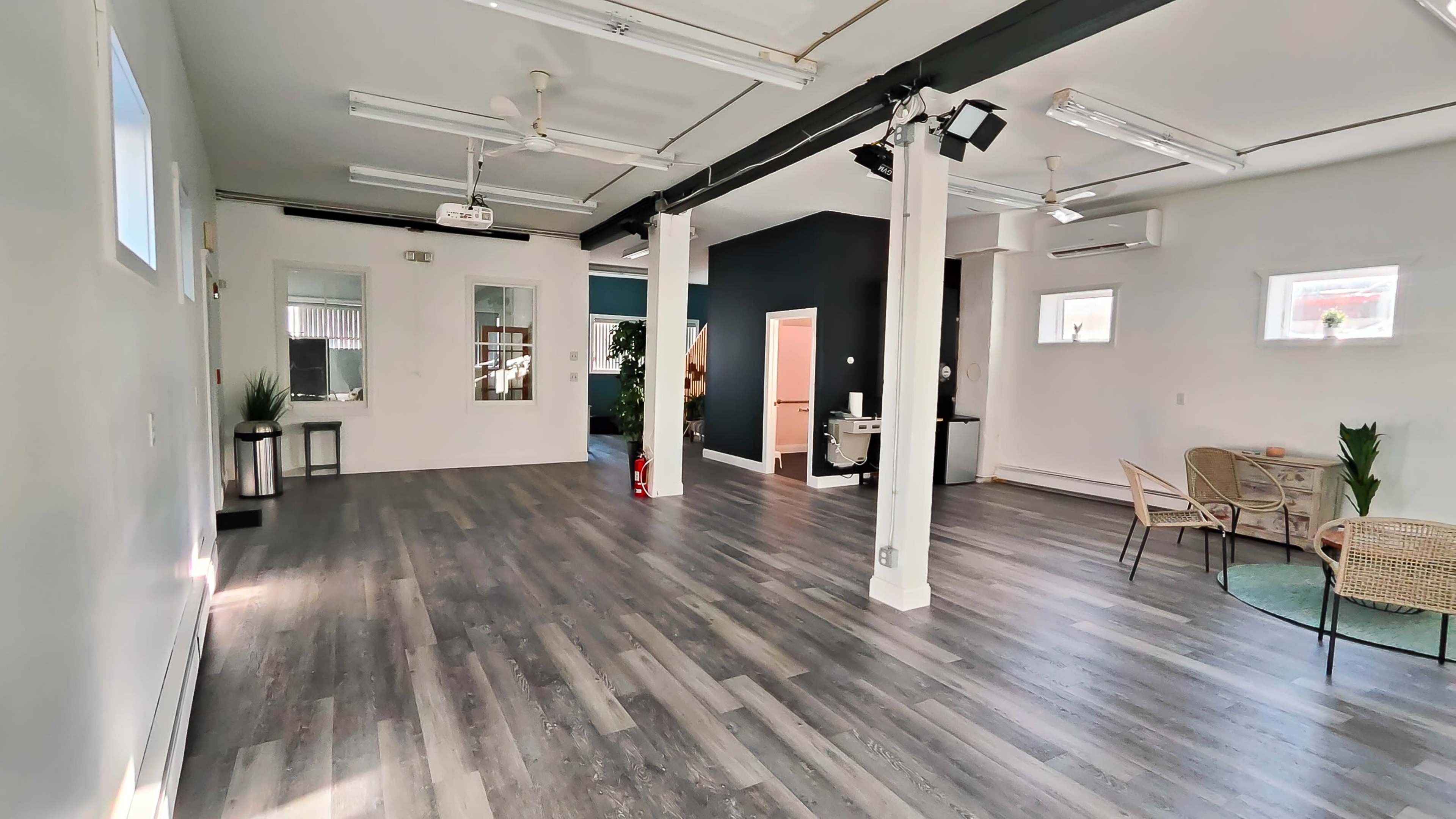 A spacious, well-lit interior with wooden floors, white walls, and a dark accent wall, featuring a few small tables and chairs.