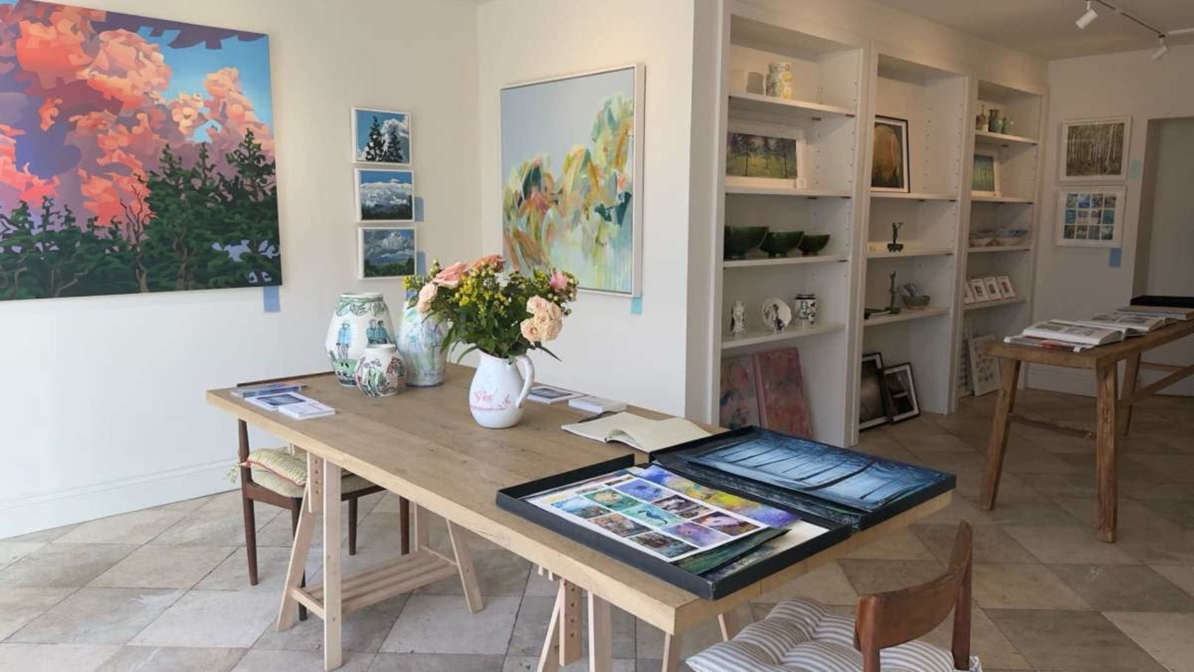 The image shows an art gallery with a wooden table displaying artwork and a vase of flowers, surrounded by shelves filled with various art pieces and photographs.