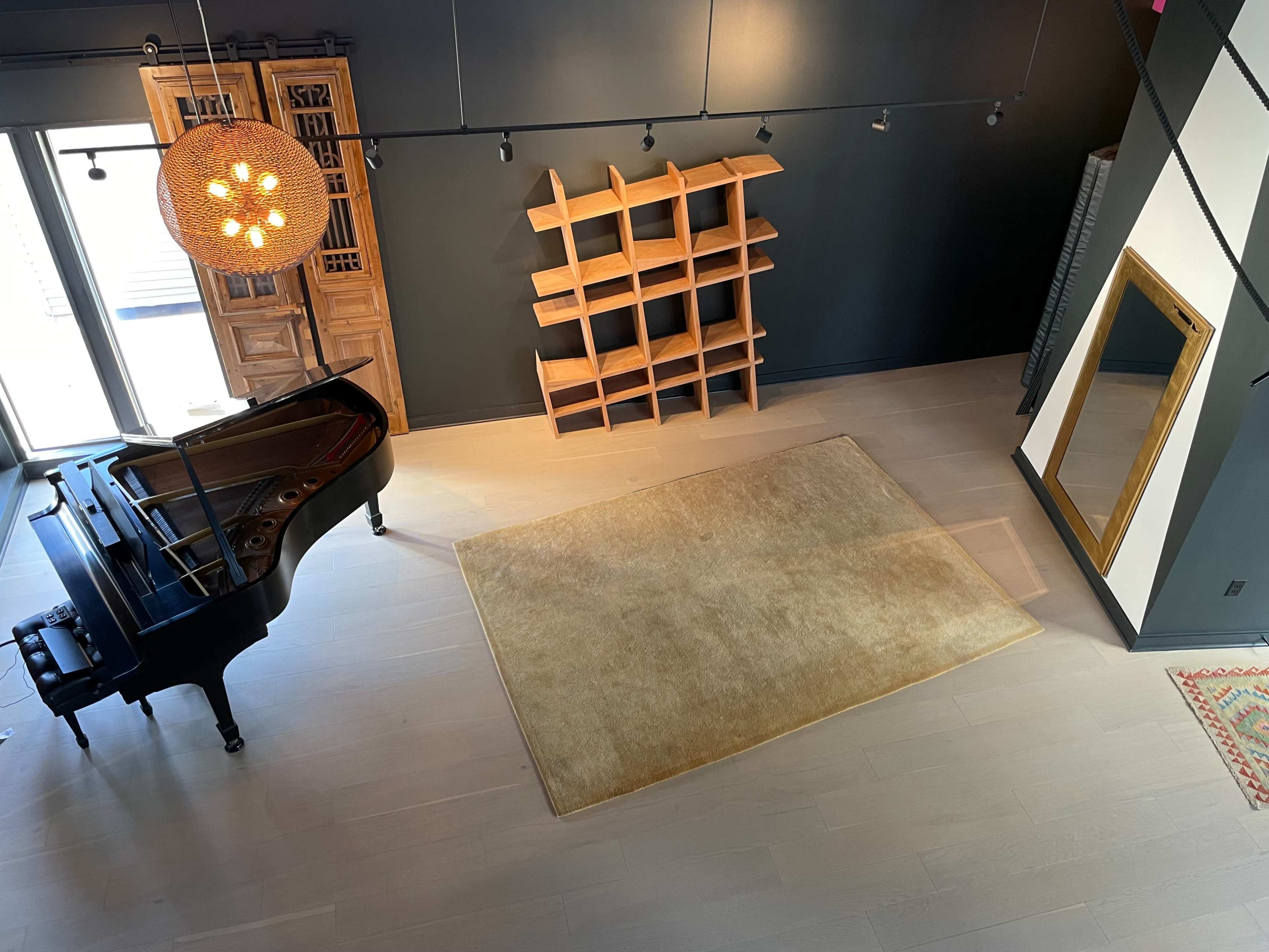 A grand piano sits beside a large rug in a spacious room featuring a wooden bookshelf and a tall mirror against a dark wall.