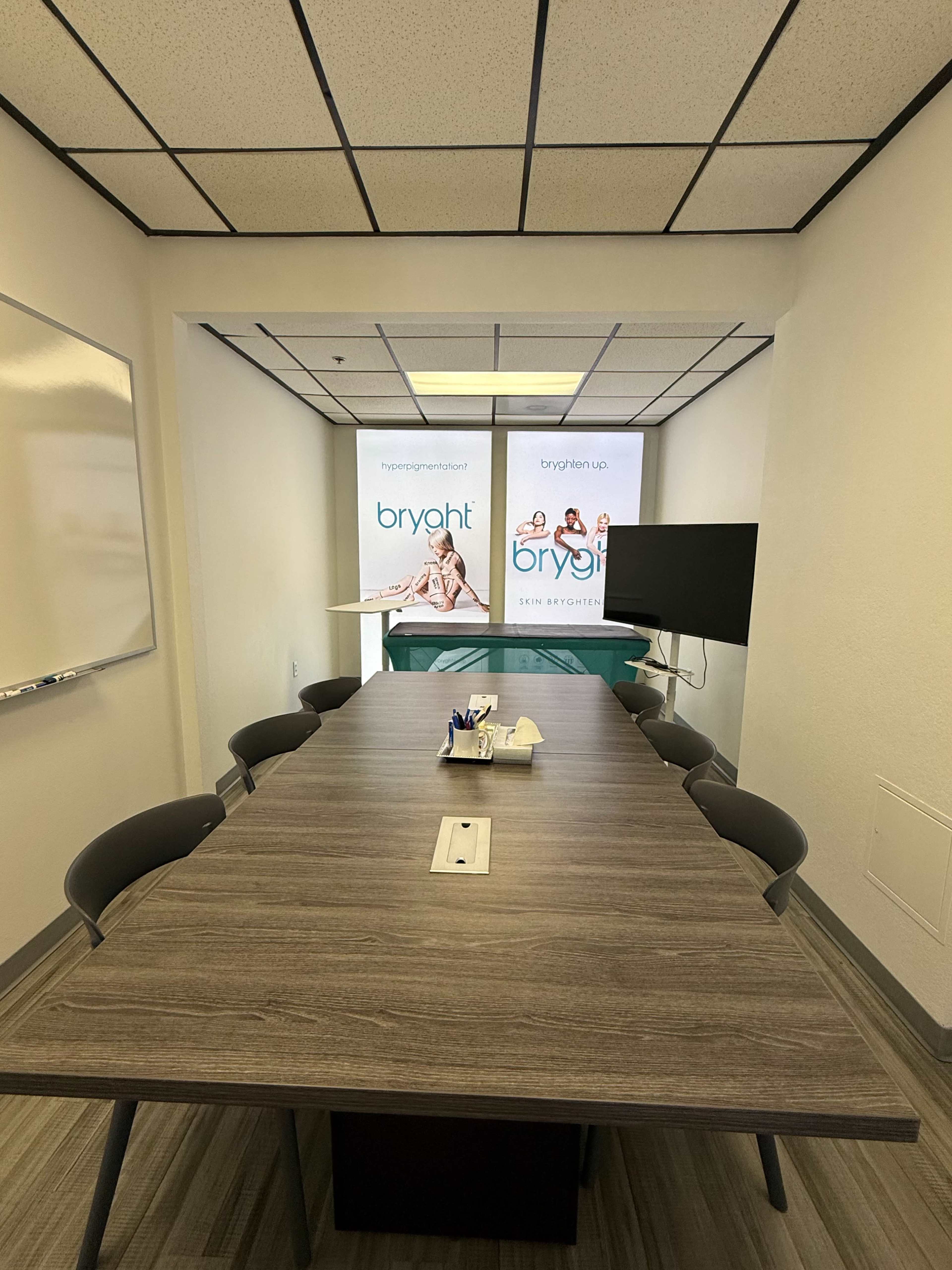 A conference room with a long wooden table, six gray chairs, a large TV mounted on the wall, and promotional banners for a brand named "bryoht" in the background.