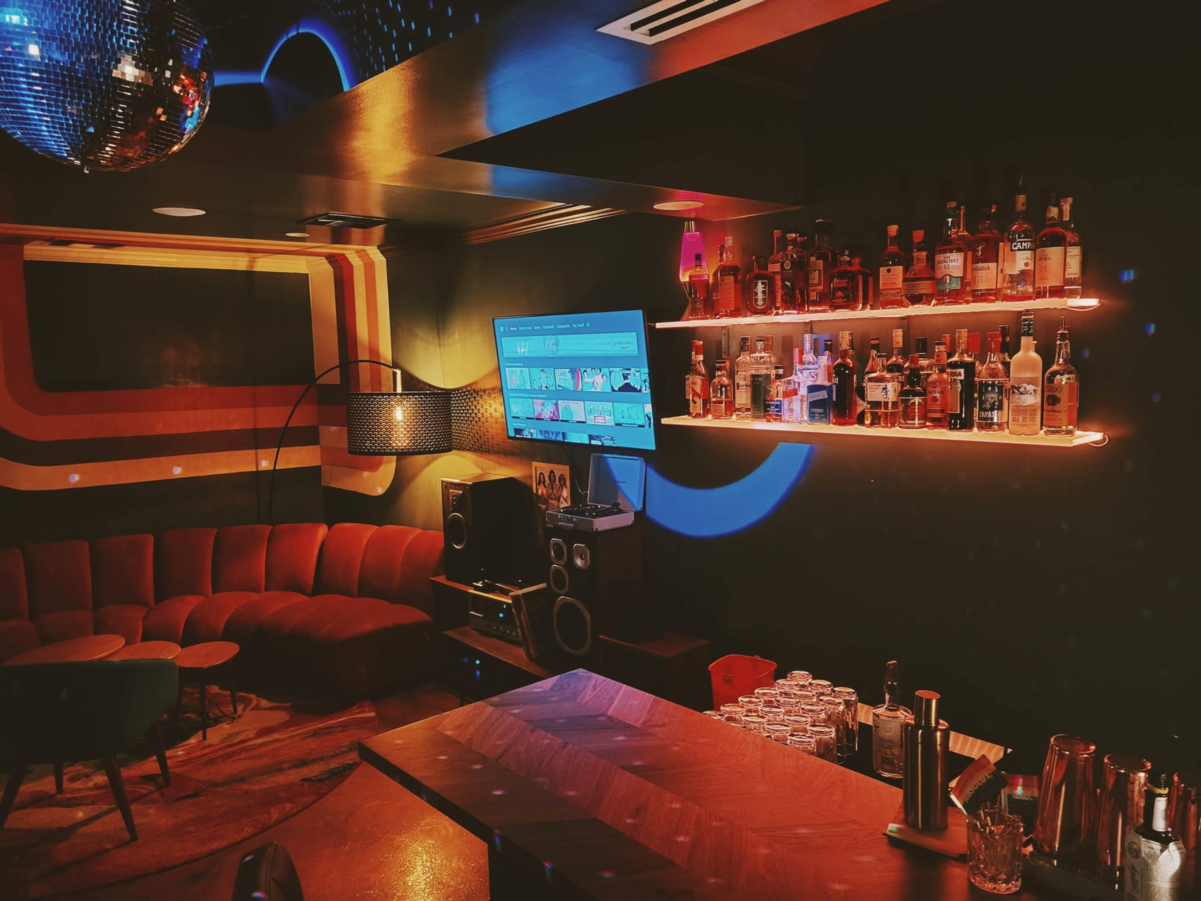 A cozy bar area features a curved red sofa, a well-stocked shelf of liquor bottles, and a large television on the wall.