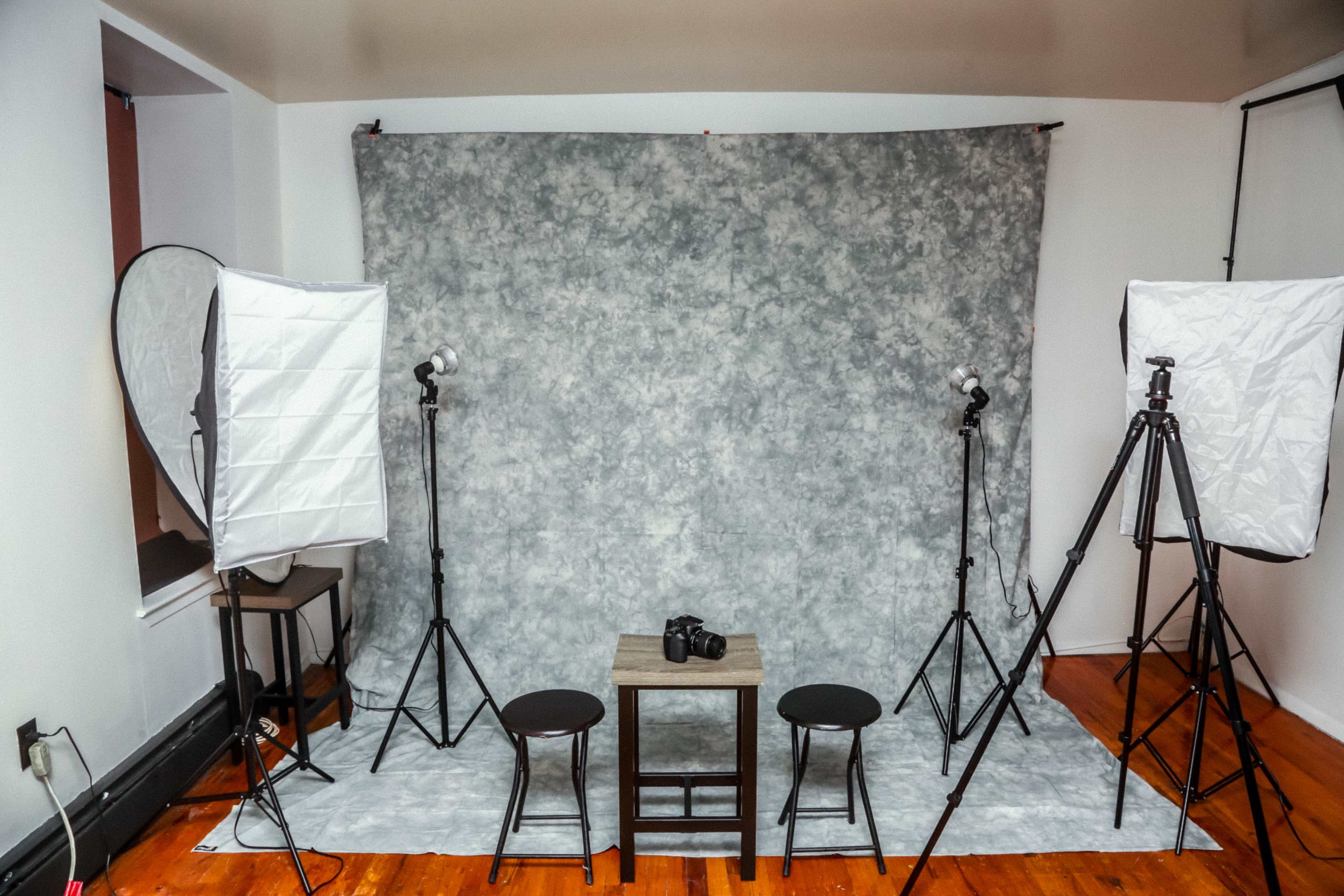 A photography setup featuring a gray backdrop, two softbox lights on tripods, and a small table with a camera positioned on it, surrounded by two stools.