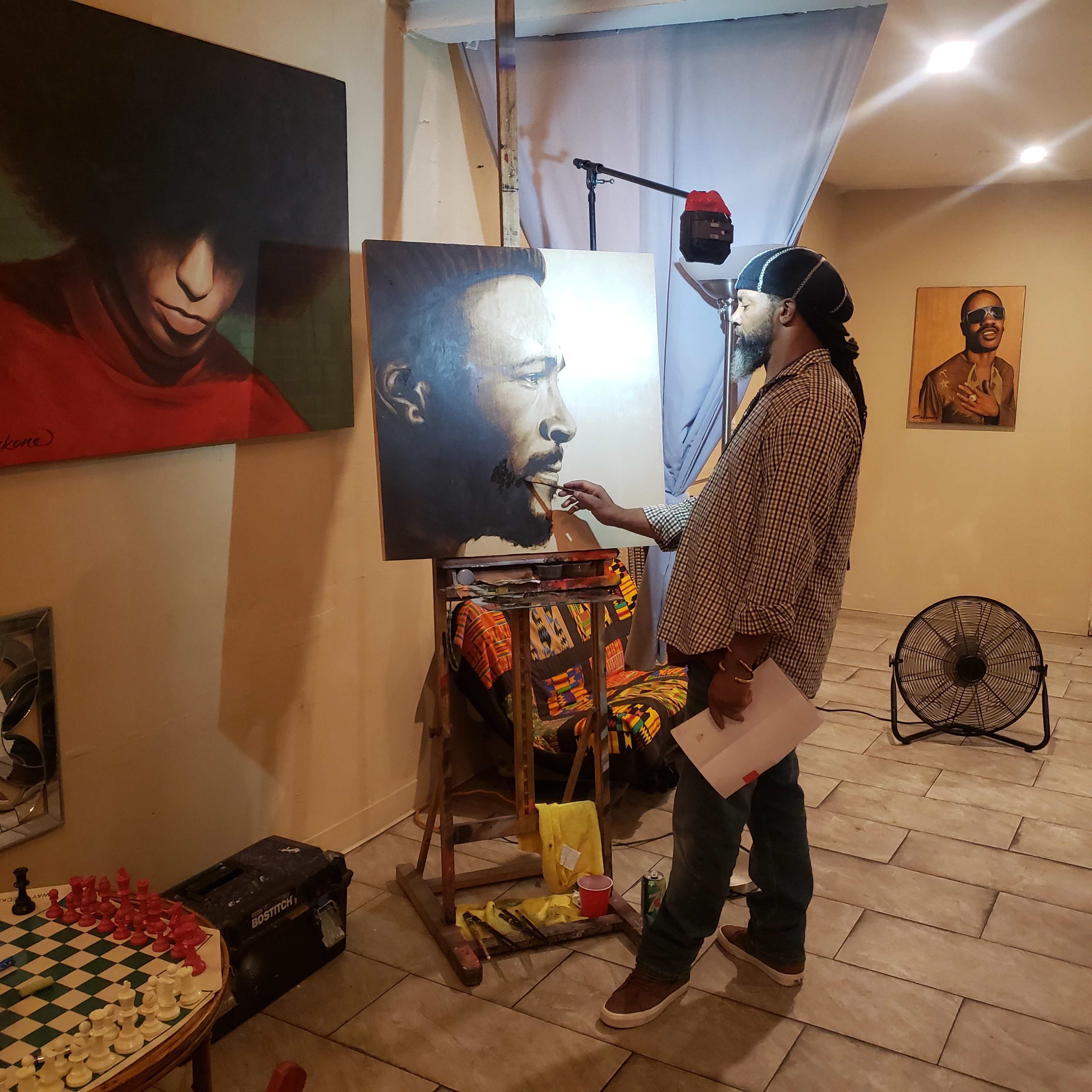 A man stands at an easel, painting a portrait of another man, while a fan and a chessboard are positioned on the floor nearby.