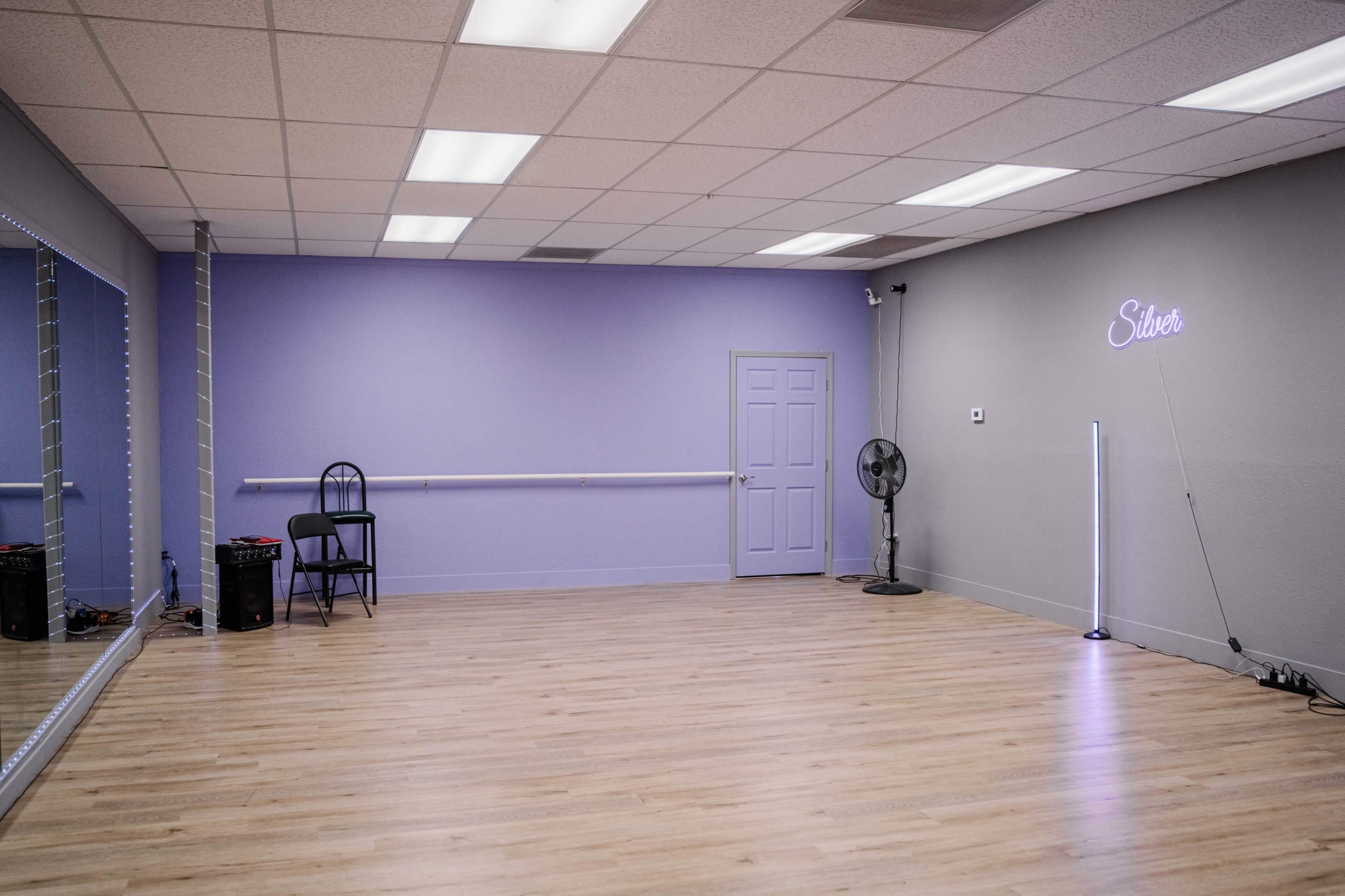 The image shows a spacious dance studio with light wooden flooring, a mirrored wall, a chair, and a fan in one corner, featuring lavender-painted walls and a neon sign that says "Silver."