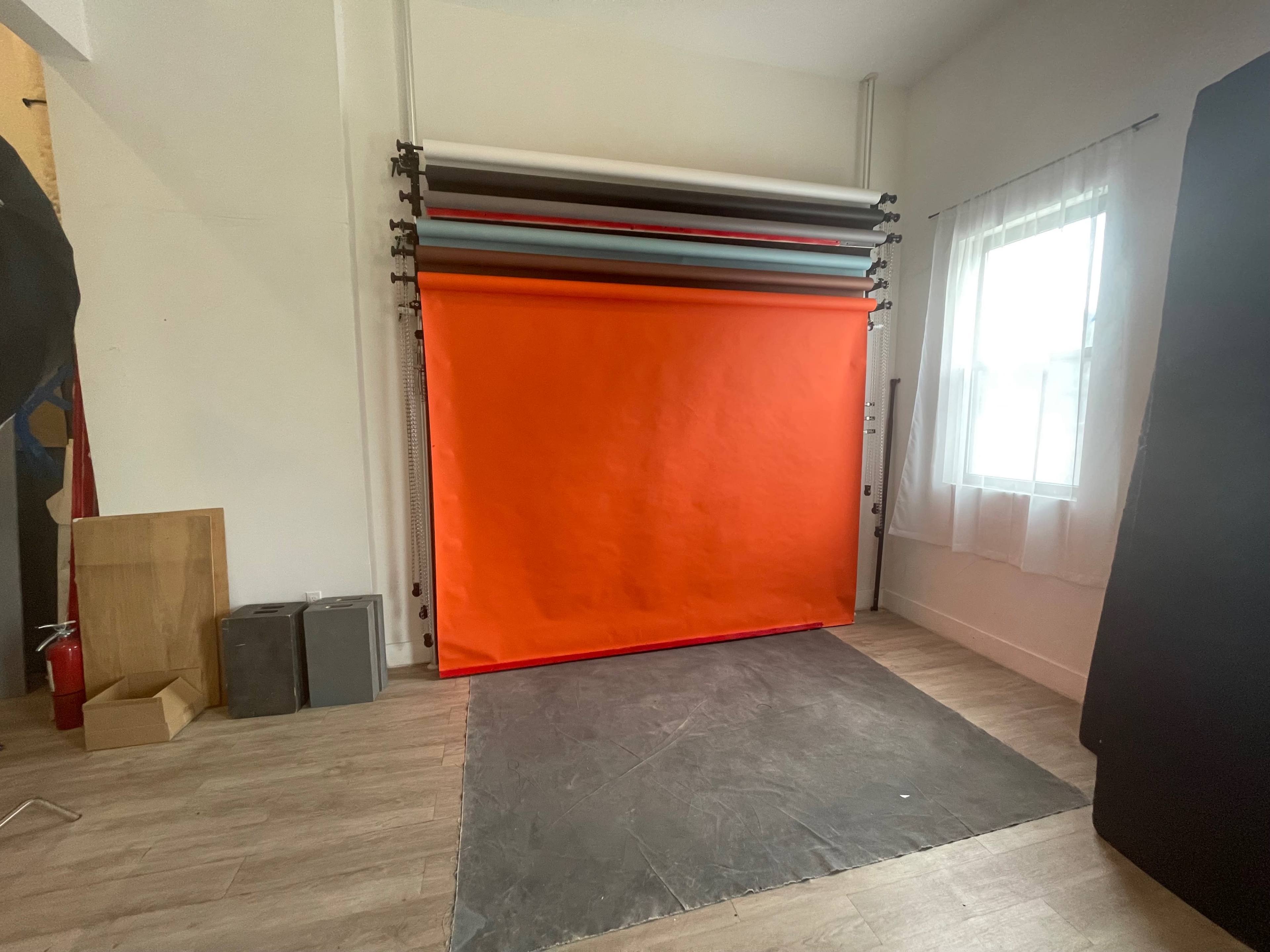 A small studio space featuring a bright orange backdrop, rolls of various colored backdrops mounted above, and a gray floor surface.