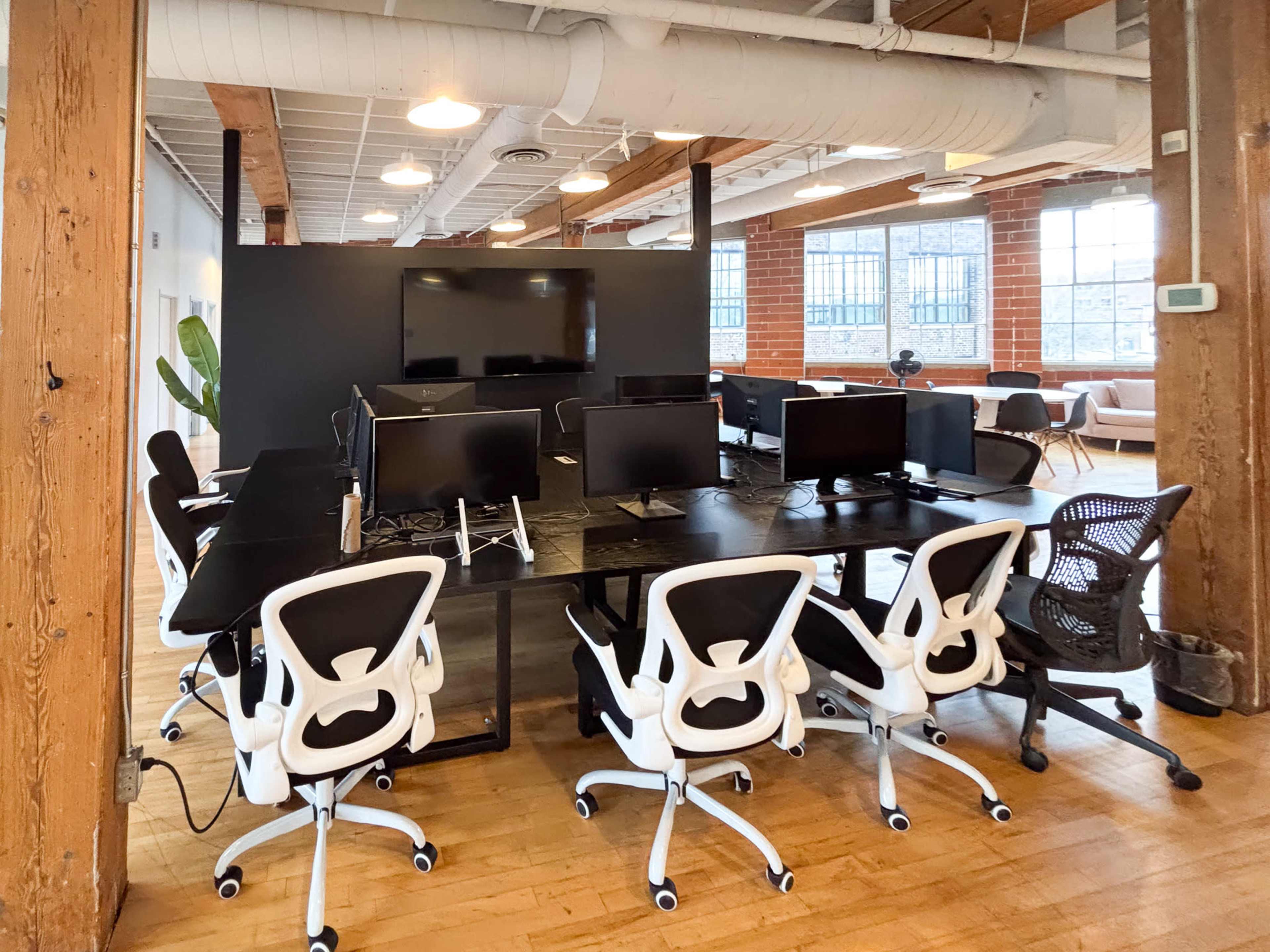 The image shows a modern office space with multiple desks, each equipped with computer monitors and ergonomic chairs, arranged in a large, open area with exposed wooden beams and large windows.