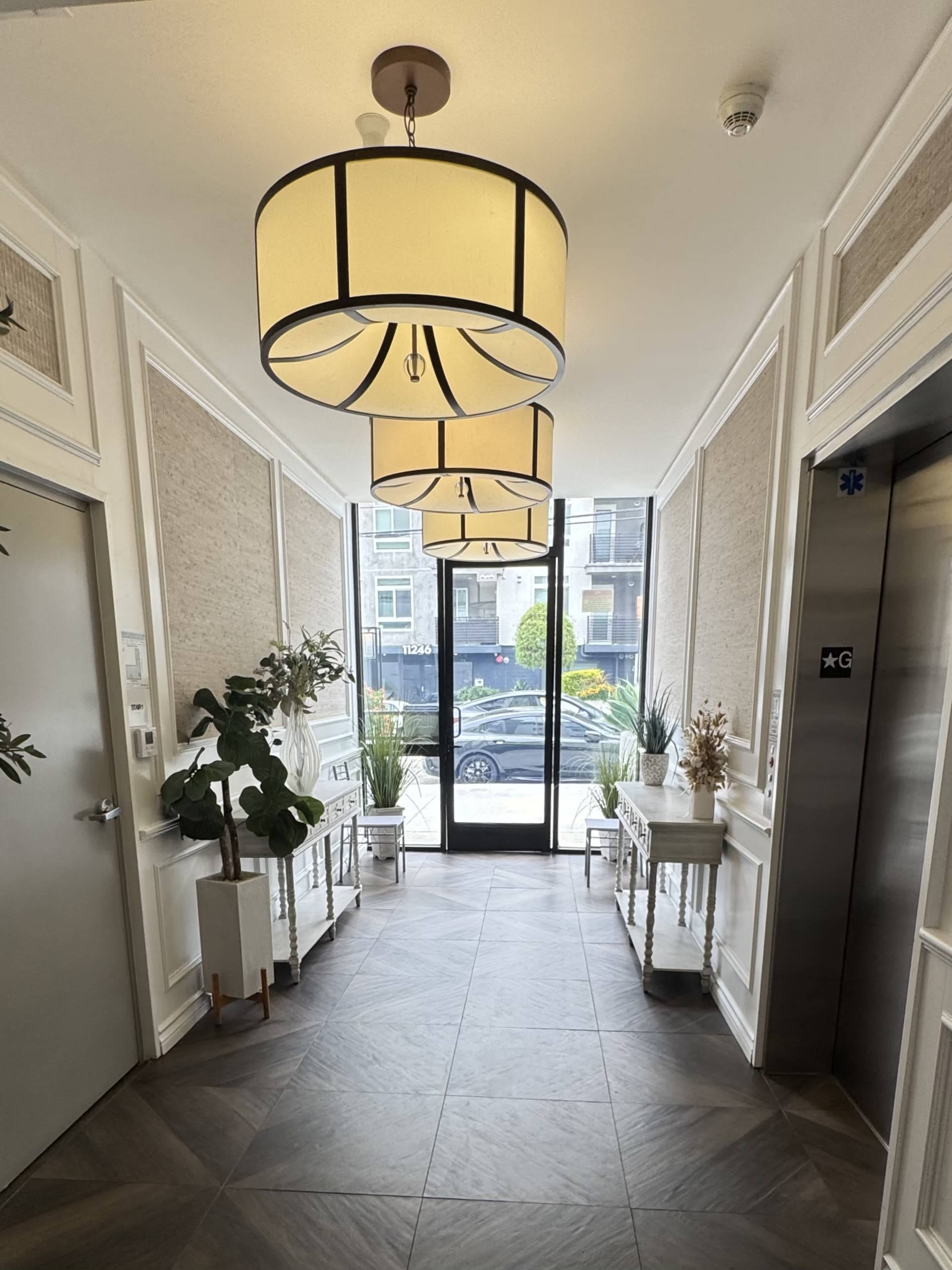 The image shows a bright hallway with three large ceiling lamps, an elevator door, potted plants, and decorative tables near a glass entrance.