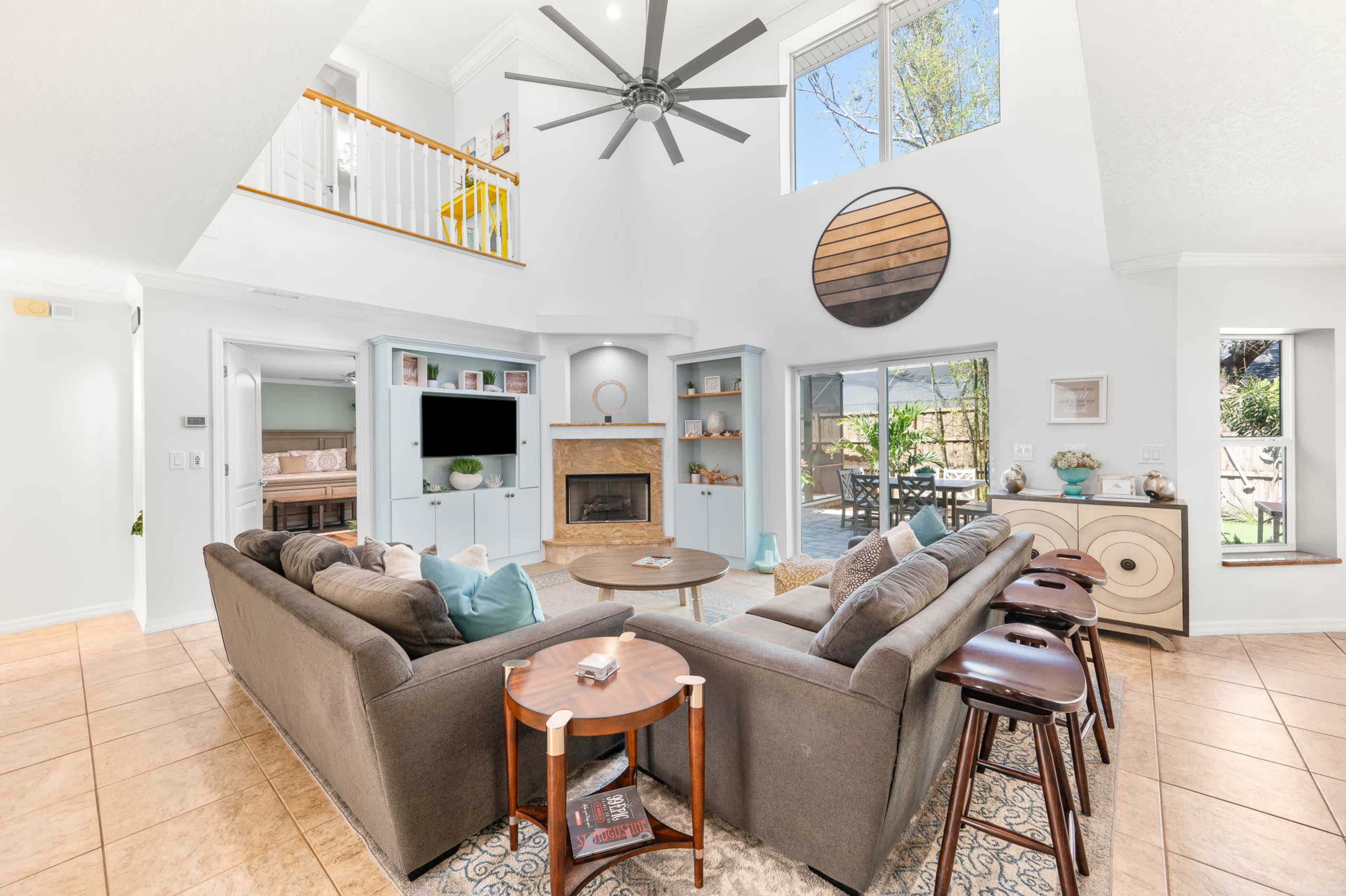 The image shows a bright and spacious living room featuring a large sofa, a coffee table, and a fireplace, with large windows allowing natural light to enter.