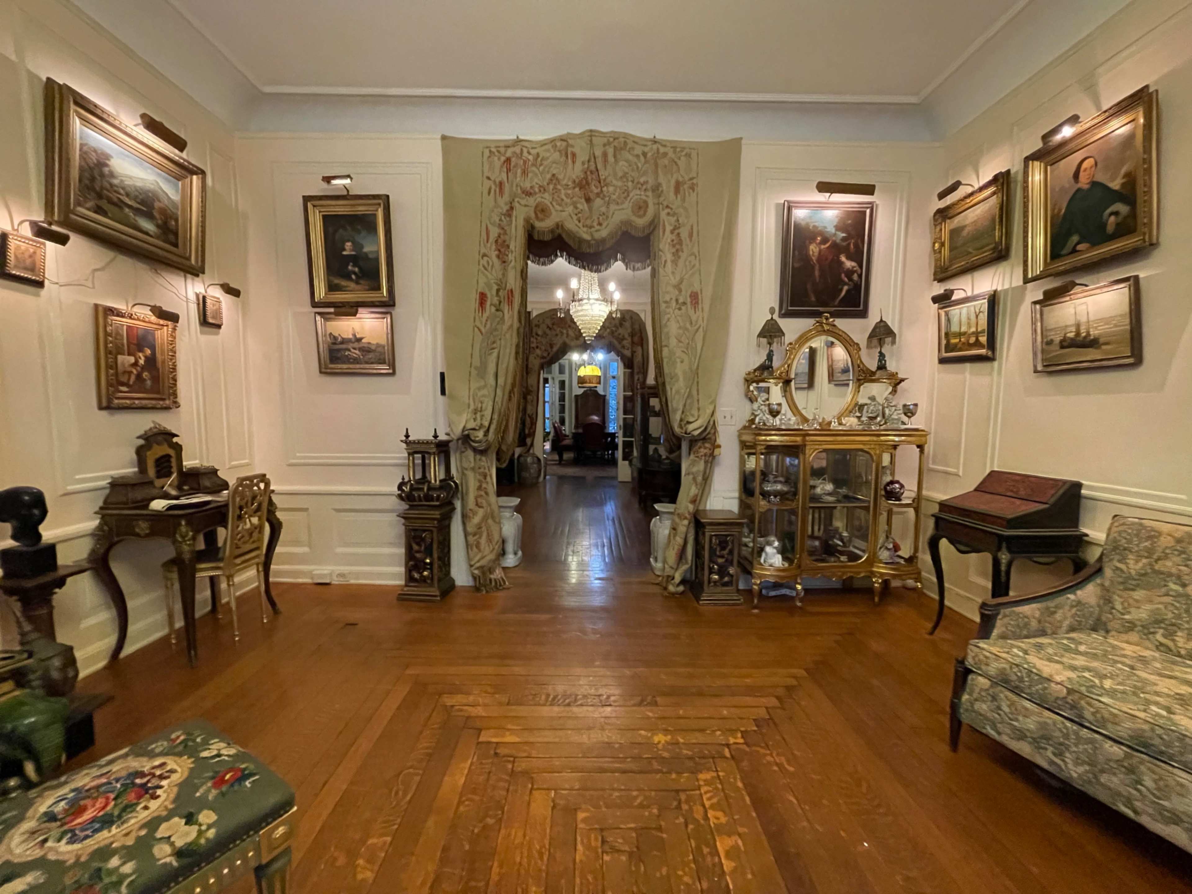 The image shows a dimly lit antique-filled room with wooden flooring, featuring framed paintings on the walls, a chandelier, and a doorway leading to another room.