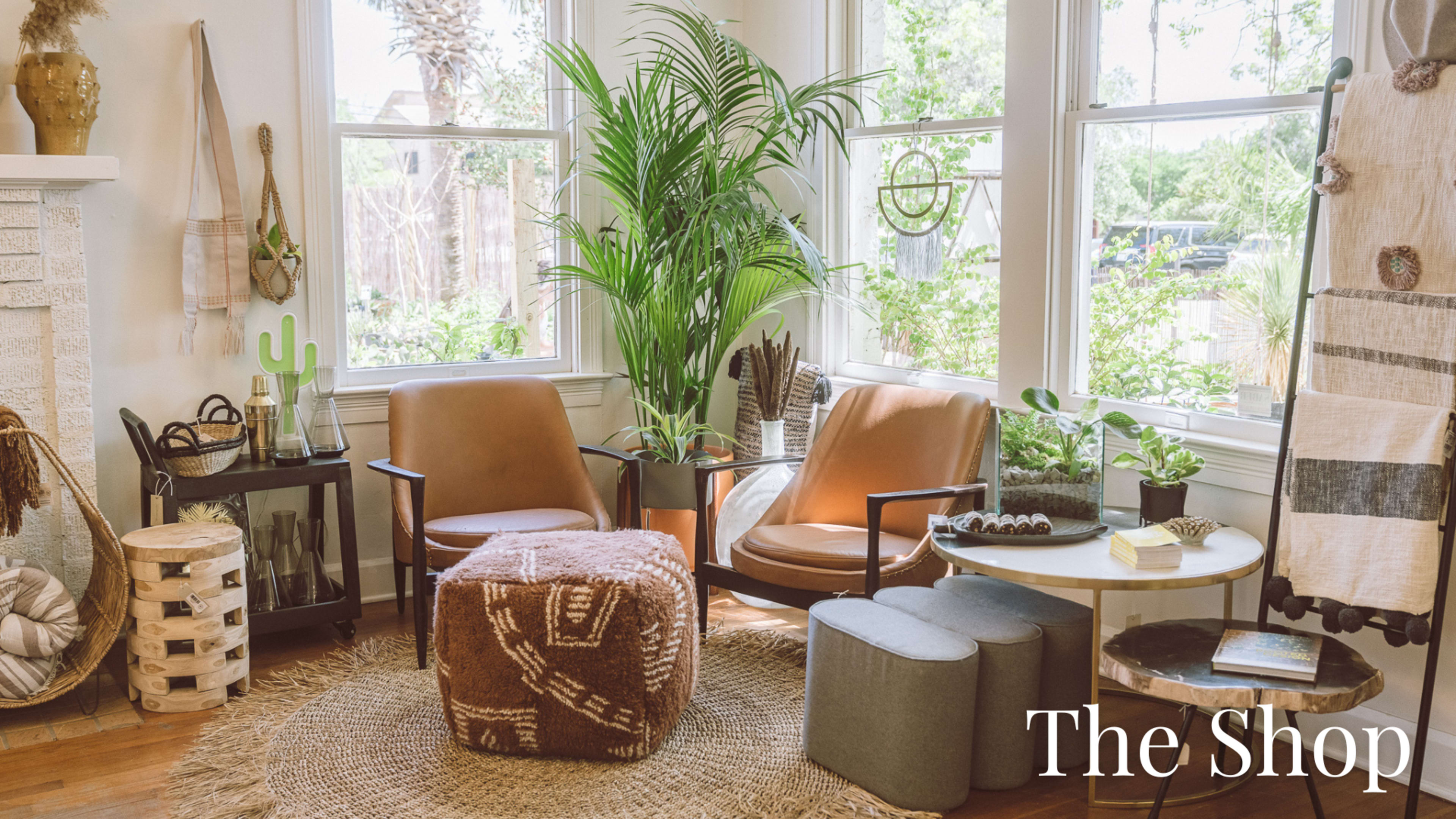 The image shows a cozy interior space with two armchairs, a round table, plants, and various decorative items arranged on a patterned rug.