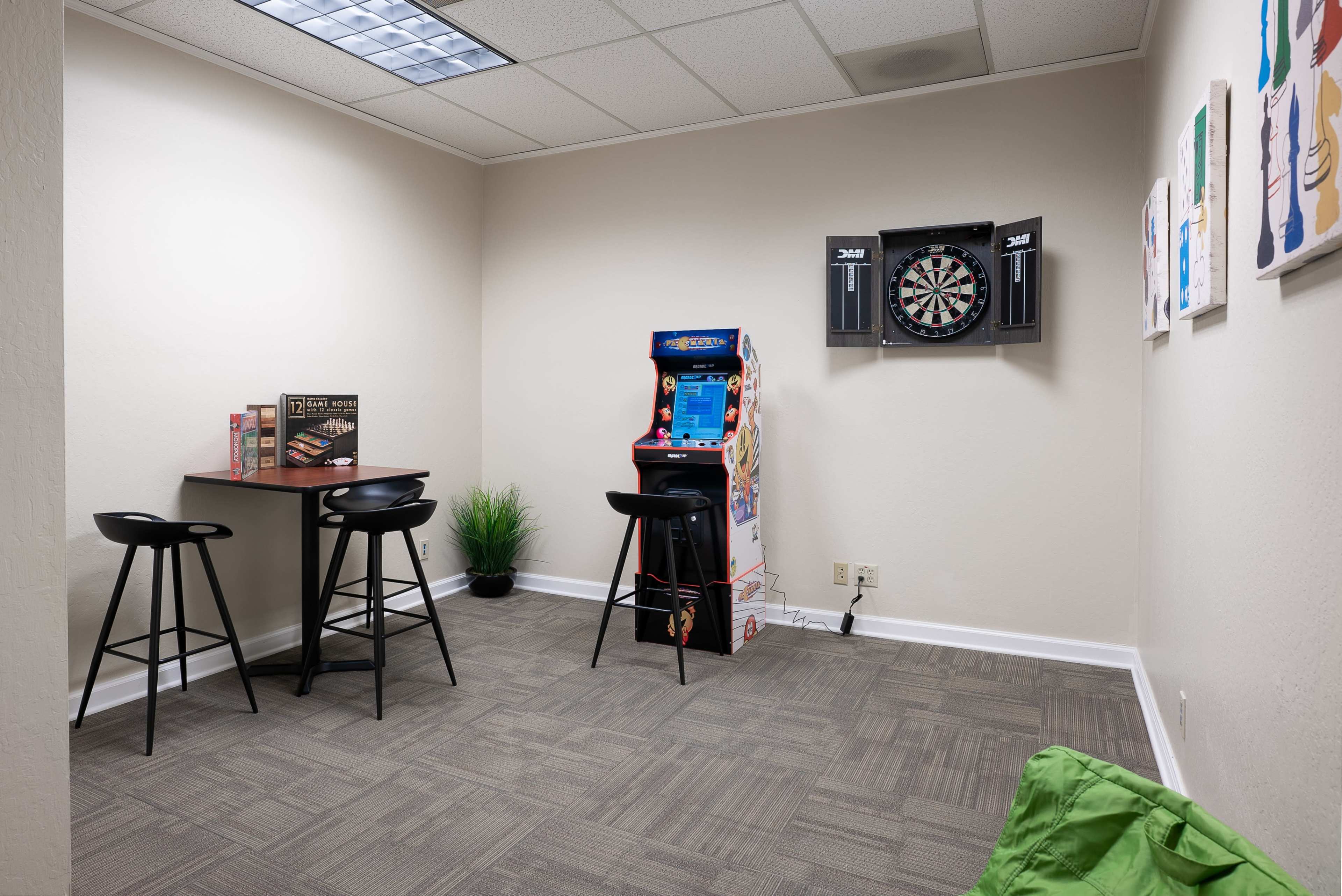 The image shows a small recreational room with a table and stools, a dartboard on the wall, an arcade machine, and green bean bags on the floor.