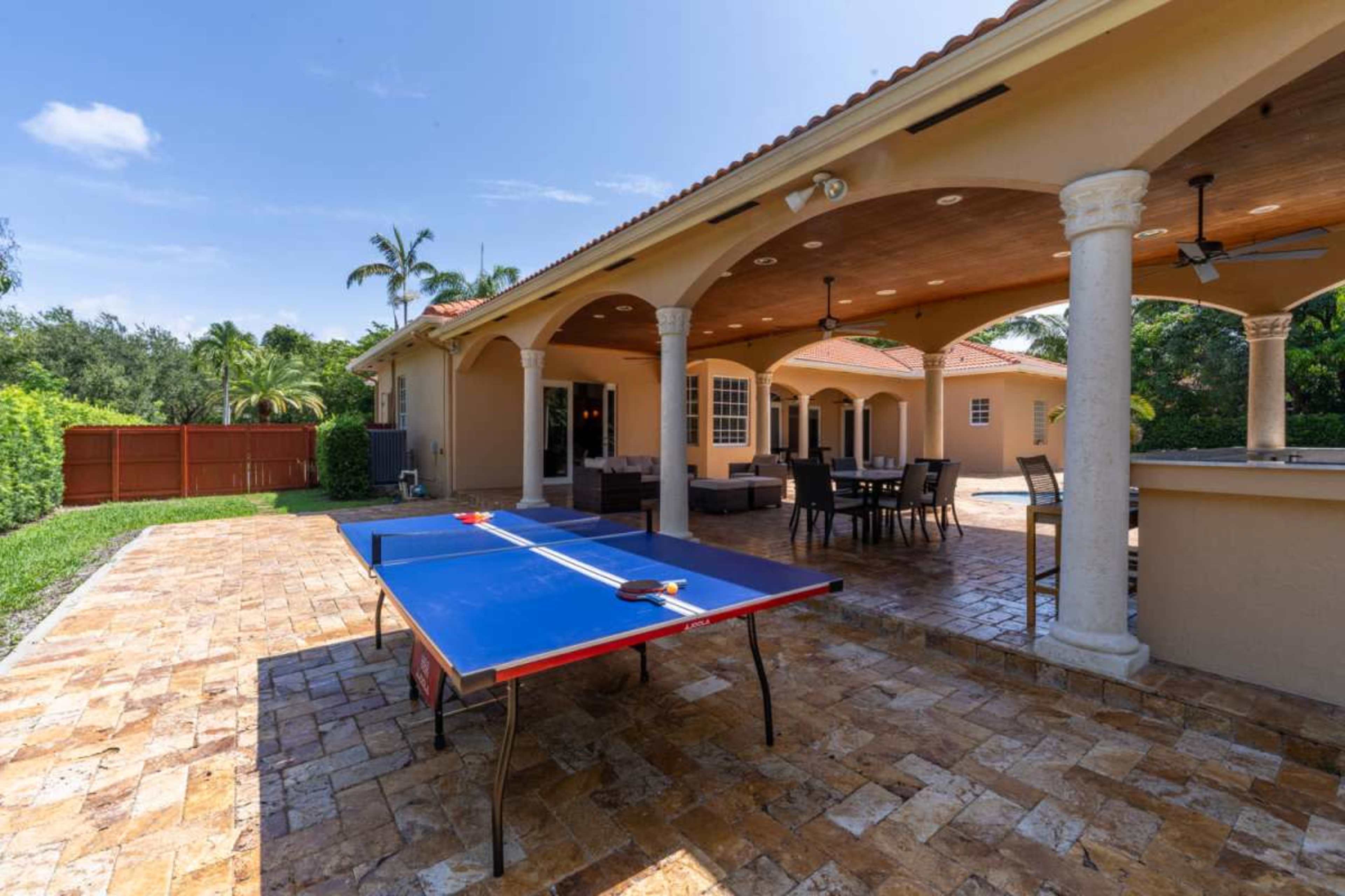 The image shows a backyard patio with a blue ping pong table and an outdoor seating area under a covered terrace.