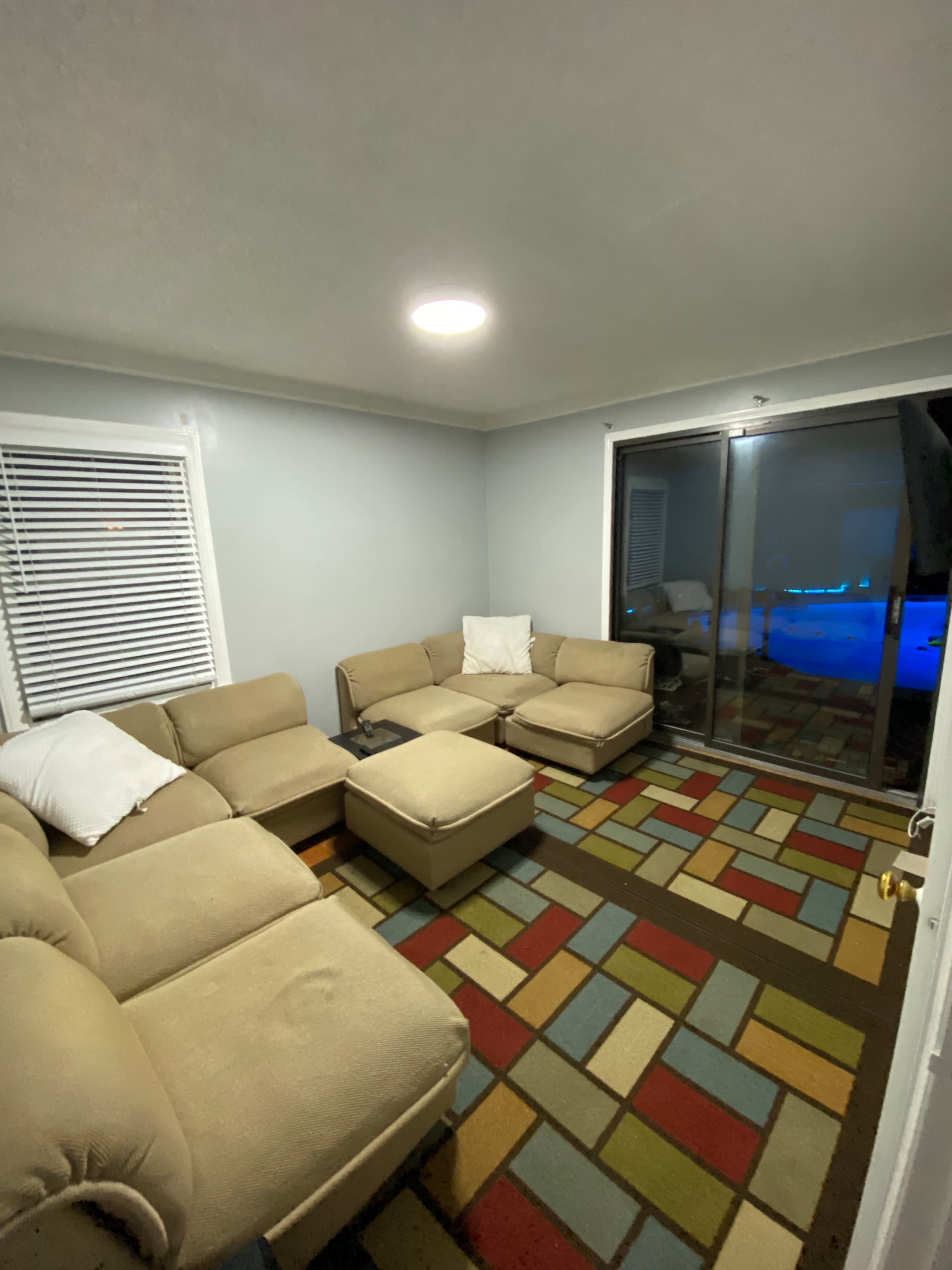 A small, dimly lit living room with a sectional sofa, an ottoman, and a colorful area rug, along with a sliding glass door that leads to an outdoor space.