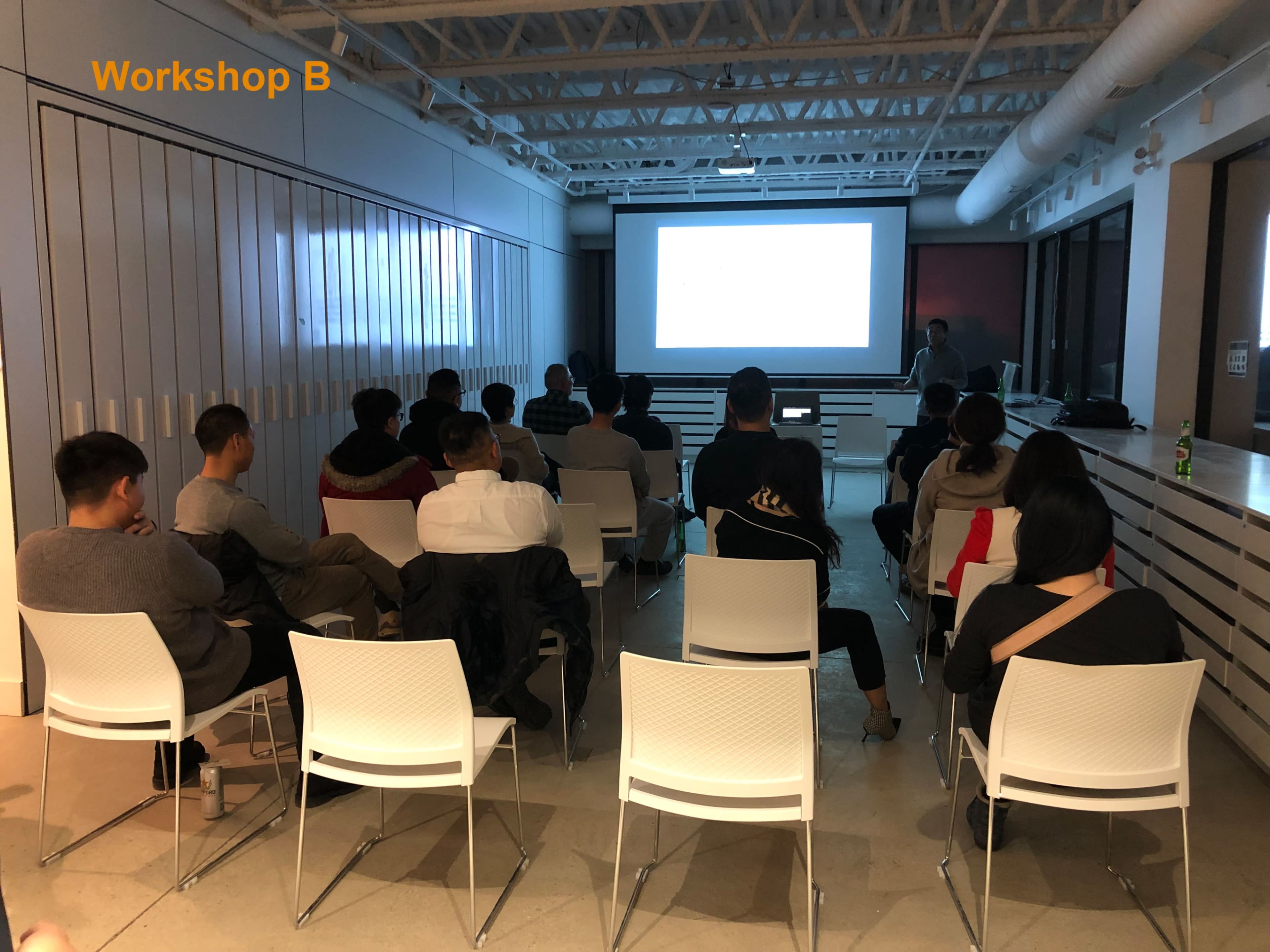 A group of people sits in white chairs facing a presentation screen in a workshop environment.