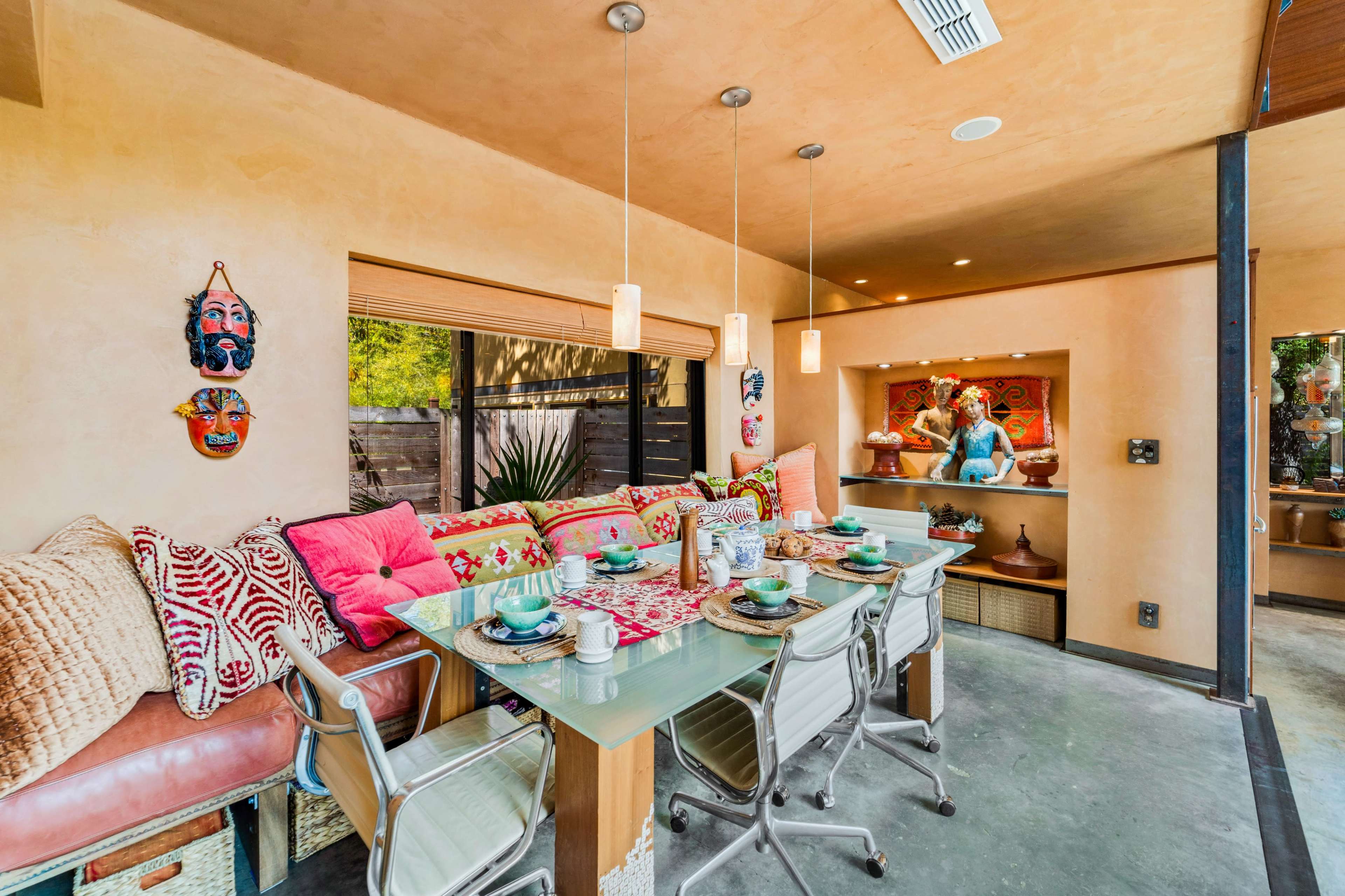 A dining area features a glass table surrounded by modern chairs, with a colorful seating area adorned with decorative pillows along one wall.