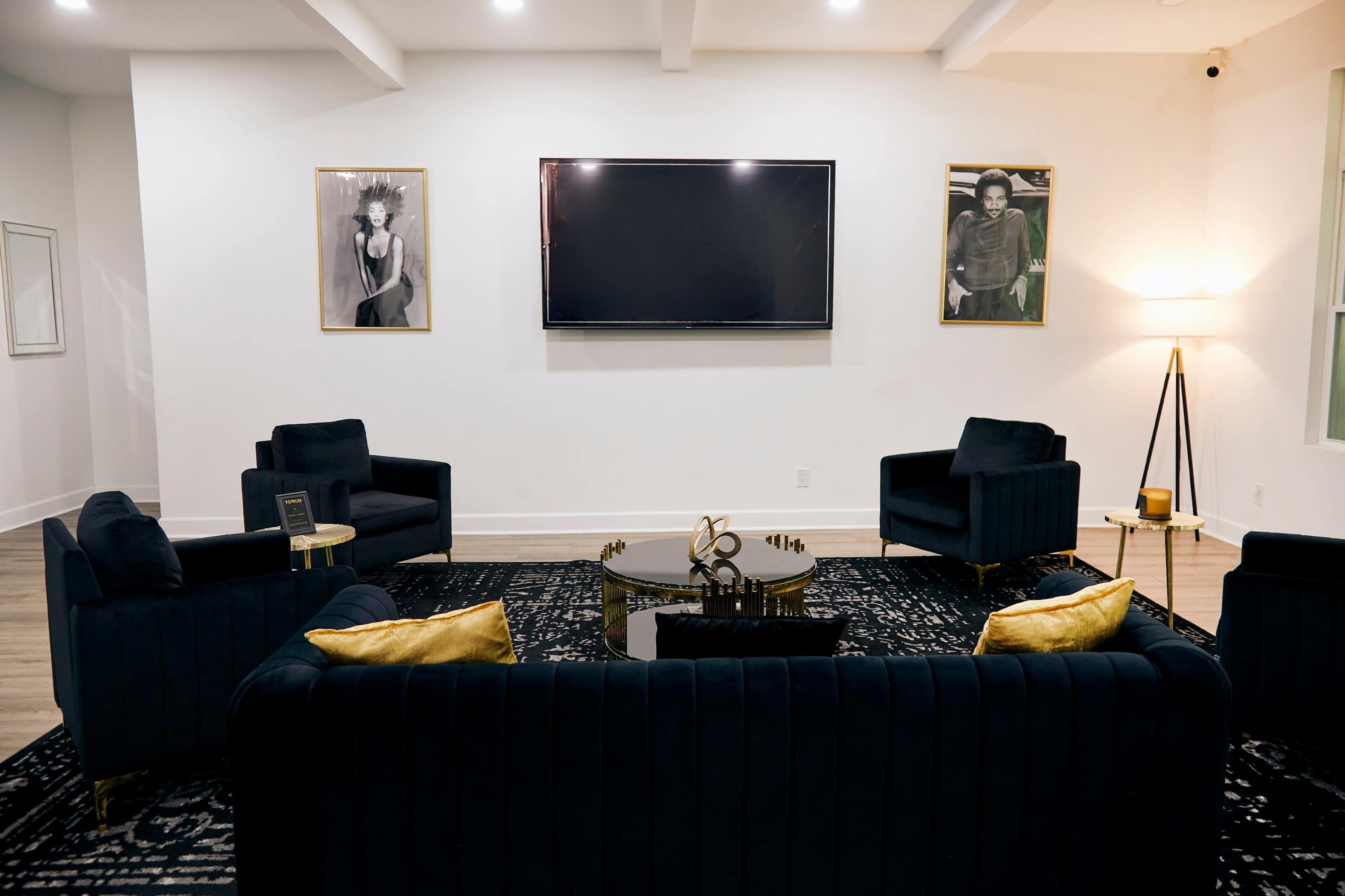 The image depicts a modern, stylish living room with dark upholstered seating, a central coffee table, and two framed portraits on the walls.