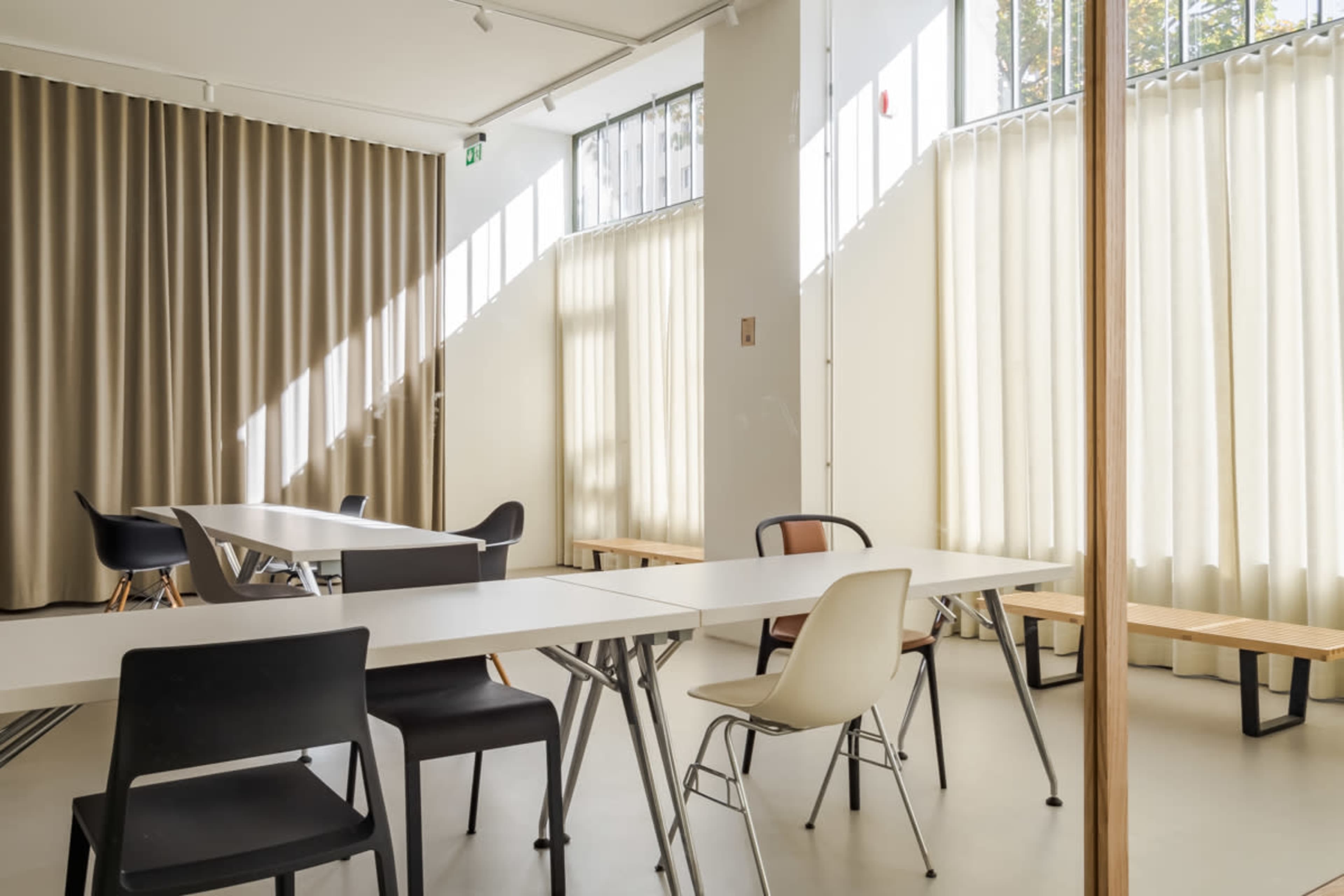 A bright, minimalist interior space with several tables and chairs arranged near large windows adorned with light-colored curtains.