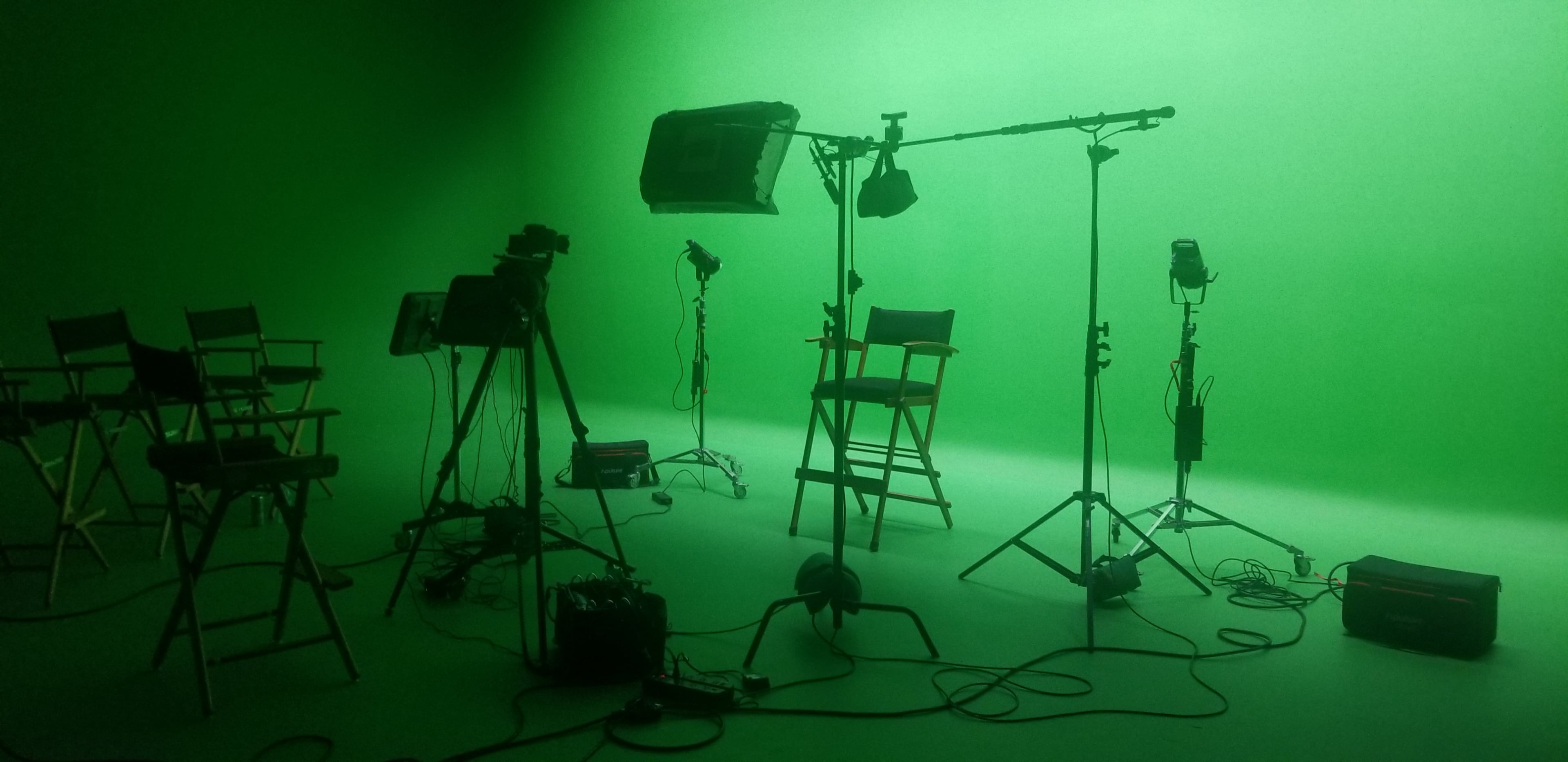 A film studio is set up with several lights, cameras, and a green backdrop, along with director's chairs arranged for a shoot.