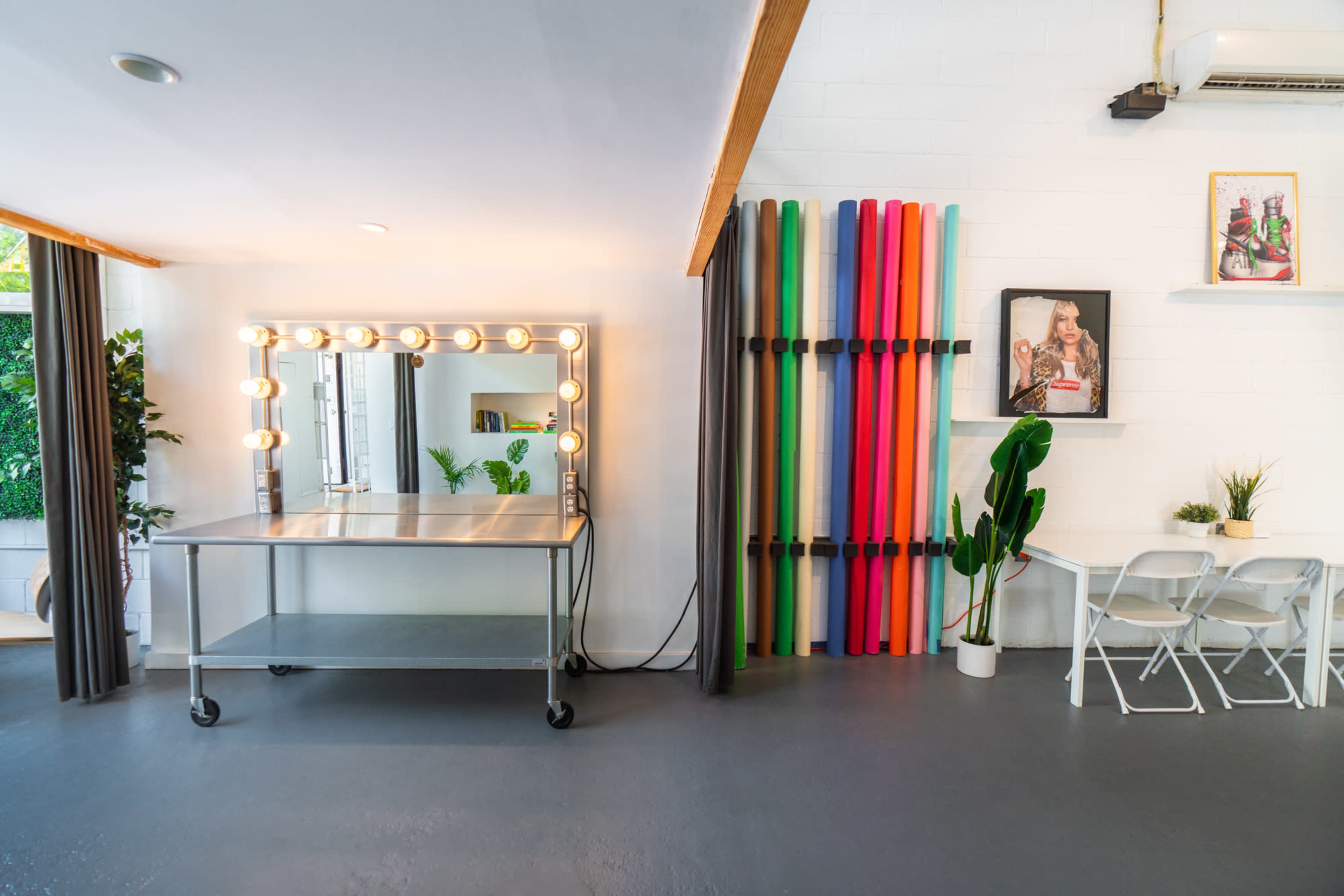 A well-lit room featuring a makeup station with a large mirror surrounded by lights, a table on wheels, colorful backdrops on a wall, and a small table with chairs.