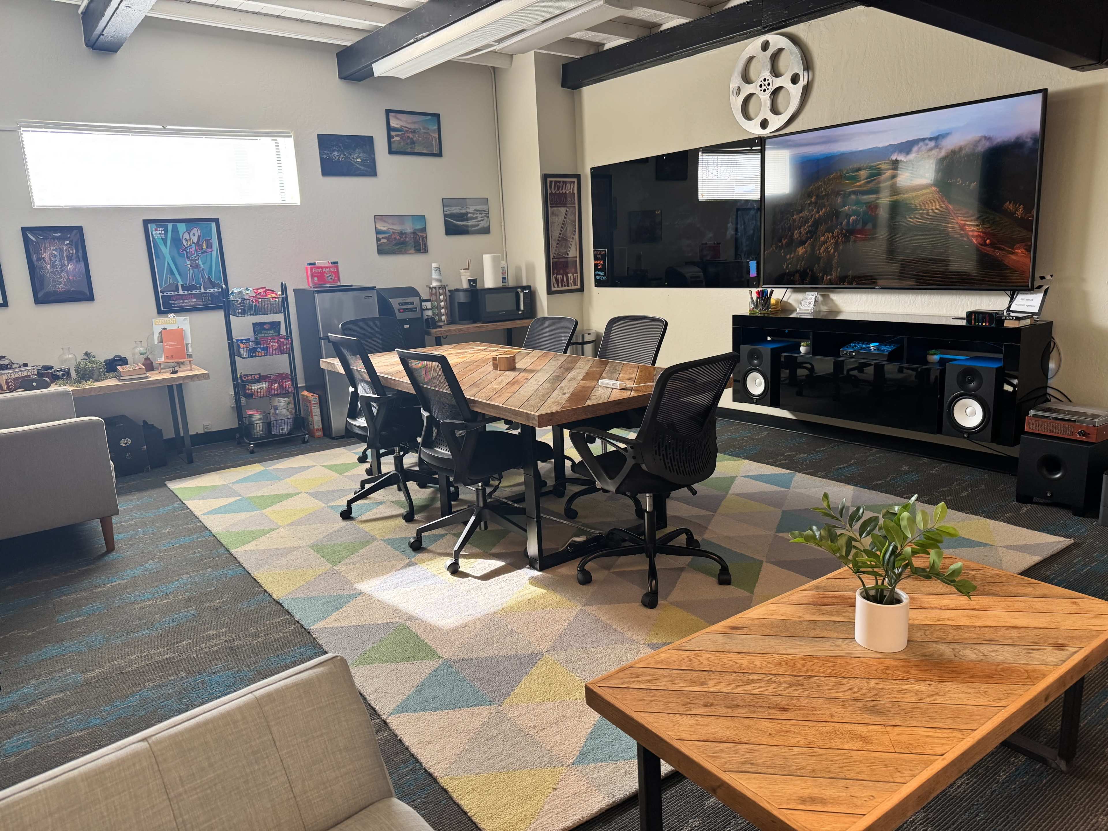 A modern meeting room features a large wooden table surrounded by ergonomic chairs, with a colorful rug on the floor and a wall-mounted screen displaying an image.