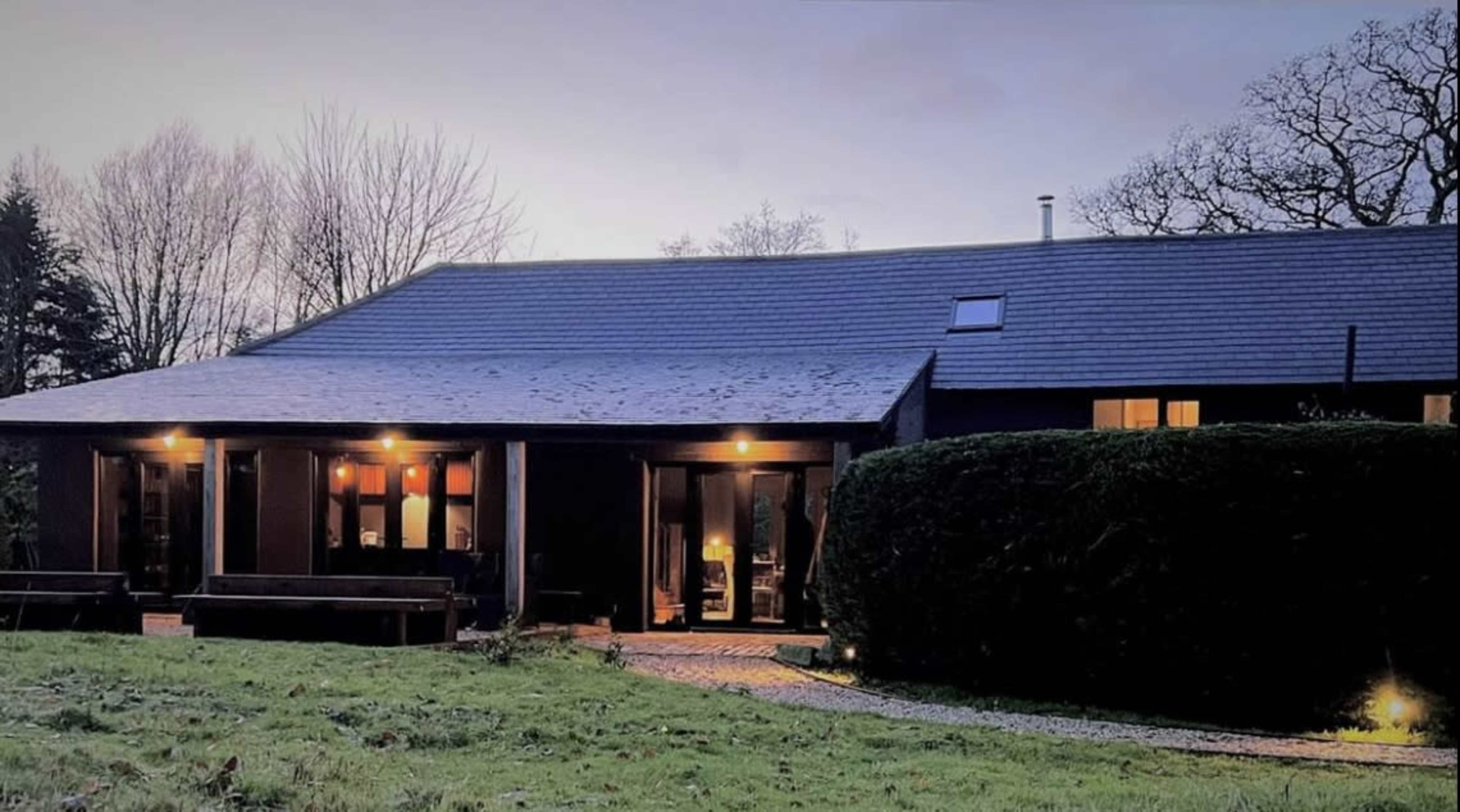 A modern house with a sloped roof and large windows sits amid a grassy area, surrounded by trees in the background.